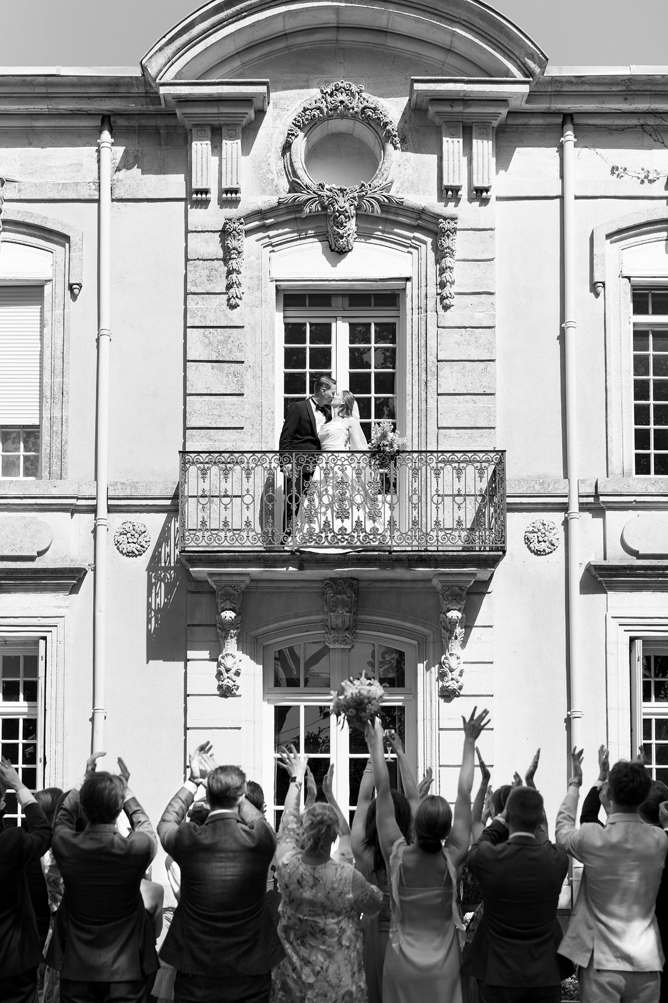 Black and white photo of a couple on a balcony celebrating their wedding with cheering guests below.