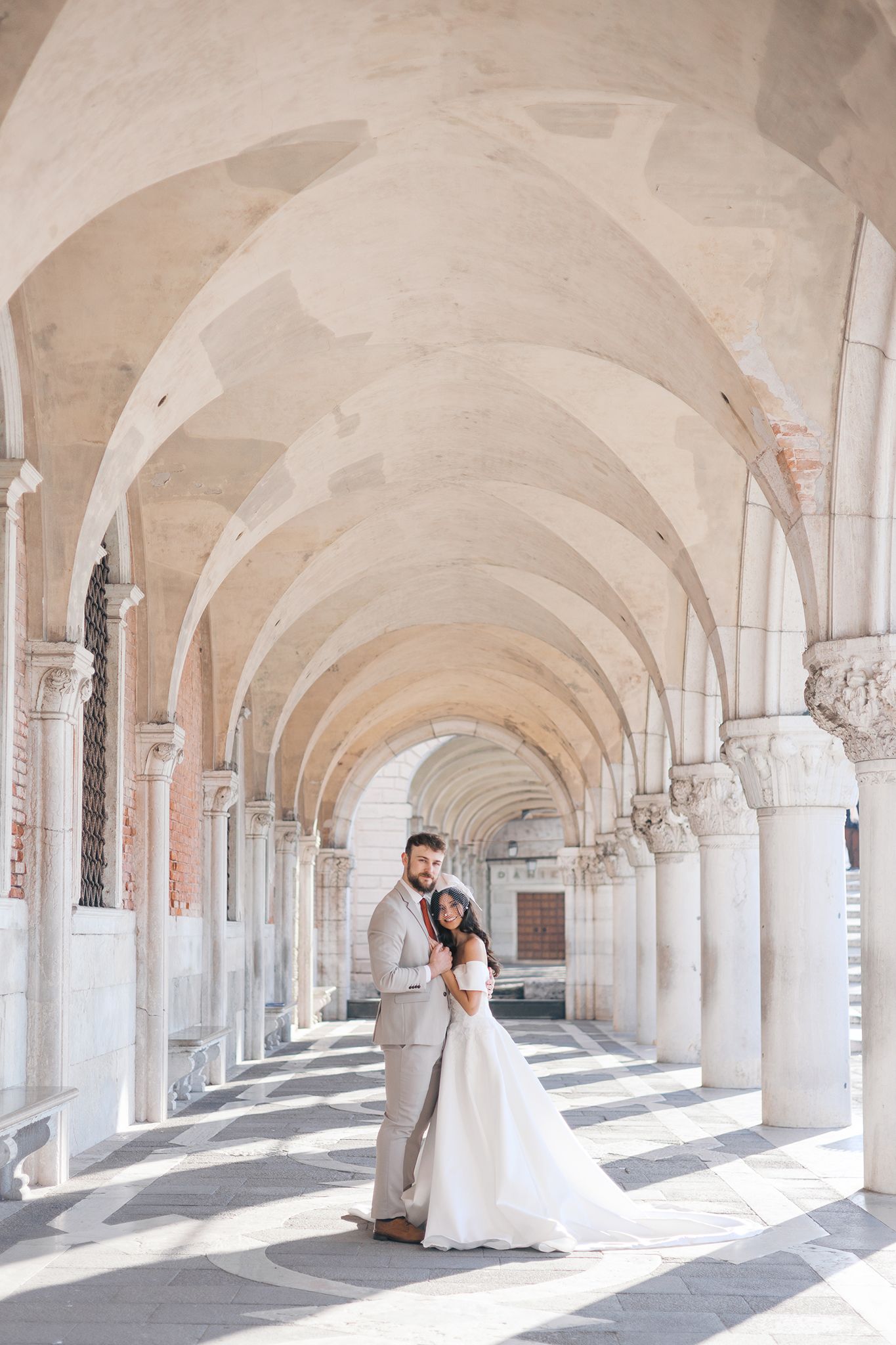 A couple embraces under beautiful arched colonnade with white stone columns and vaulted ceilings in Venice, Italy.