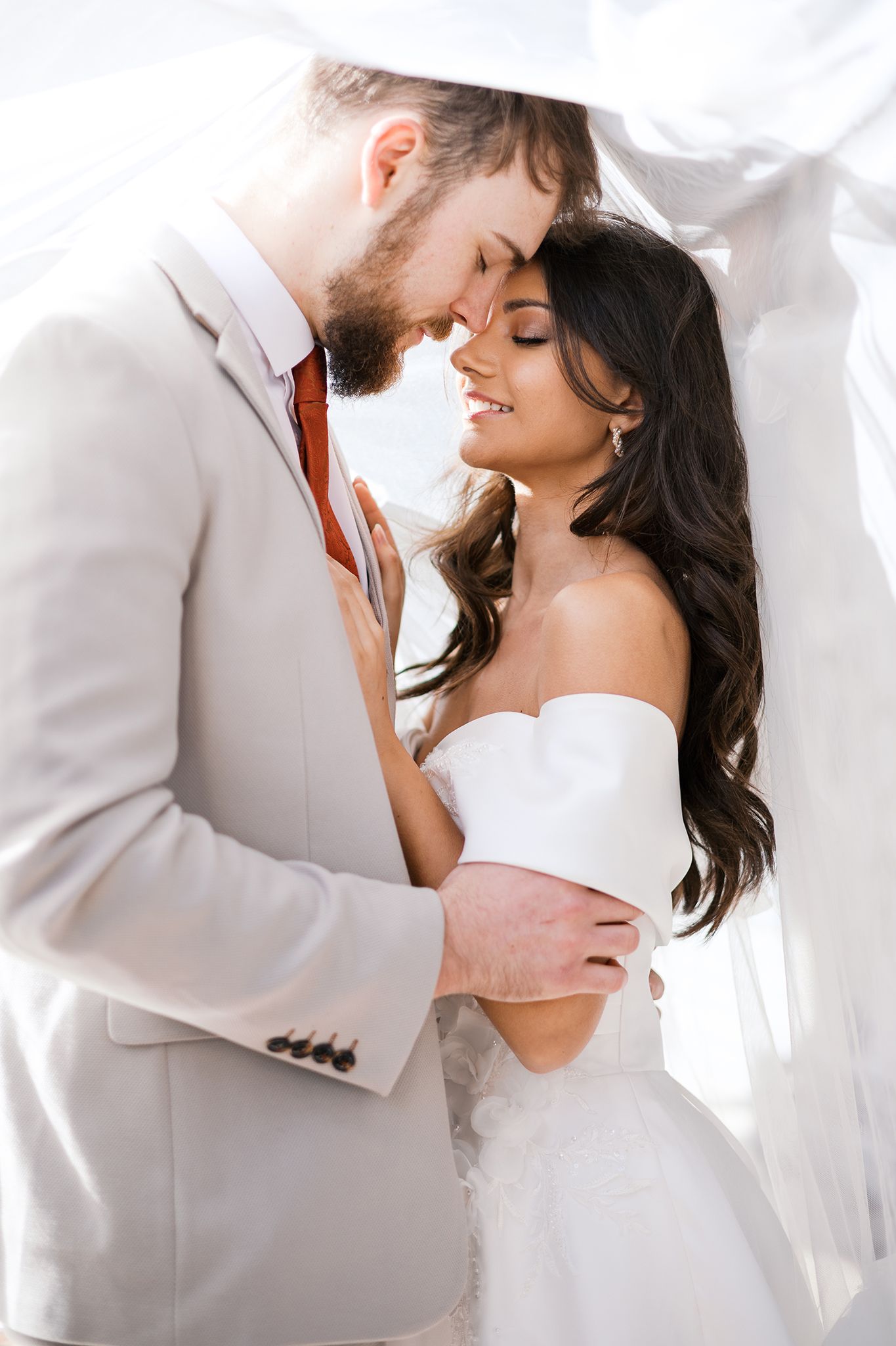 A romantic couple shares an intimate moment in white wedding attire under sheer fabric.