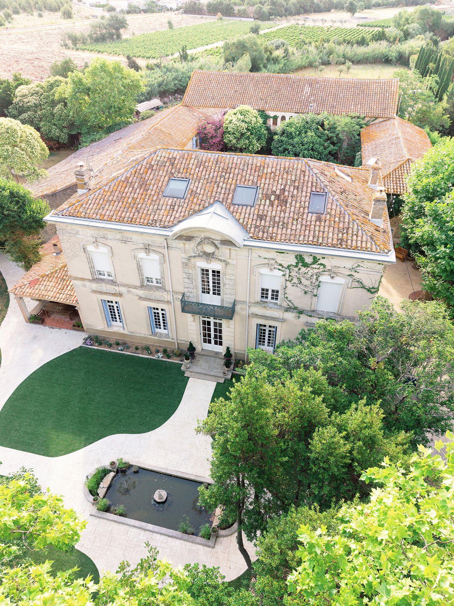 Aerial view of a grand French château with terracotta roof, manicured lawn, fountain courtyard, surrounded by lush greenery.