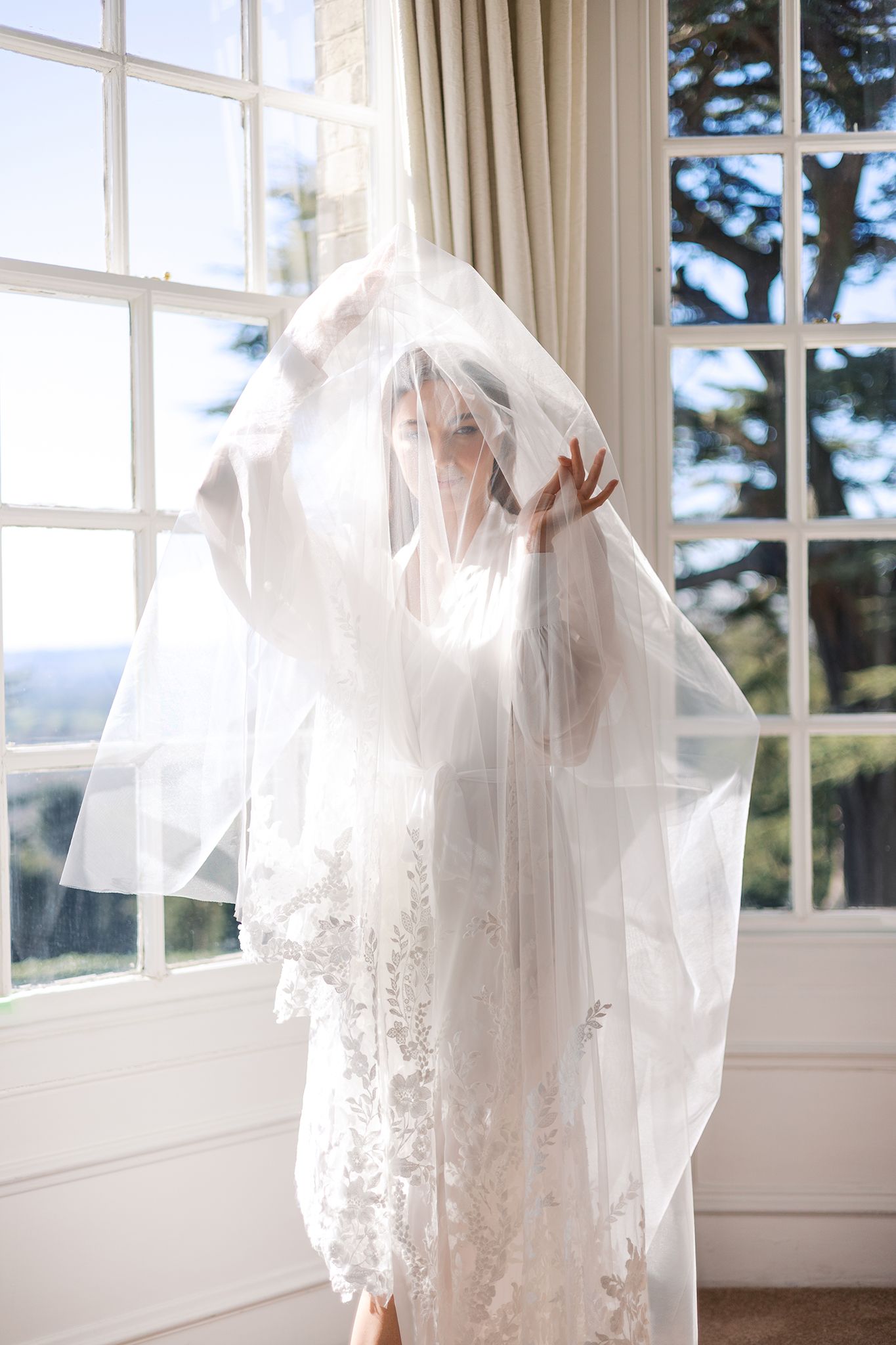 A ethereal white wedding veil and lace dress catch sunlight streaming through large windows in an elegant room.