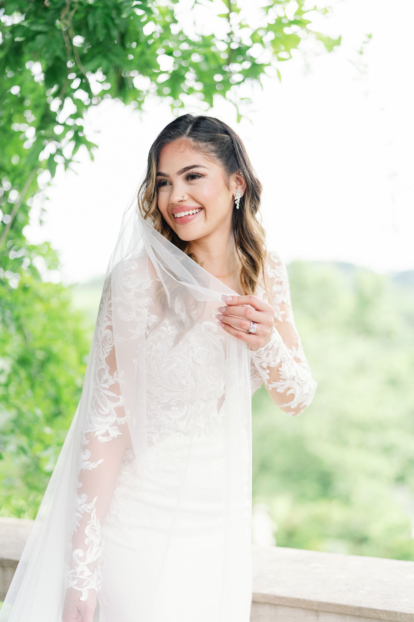 Smiling bride in lace long-sleeve wedding gown and veil poses on outdoor balcony surrounded by lush greenery.