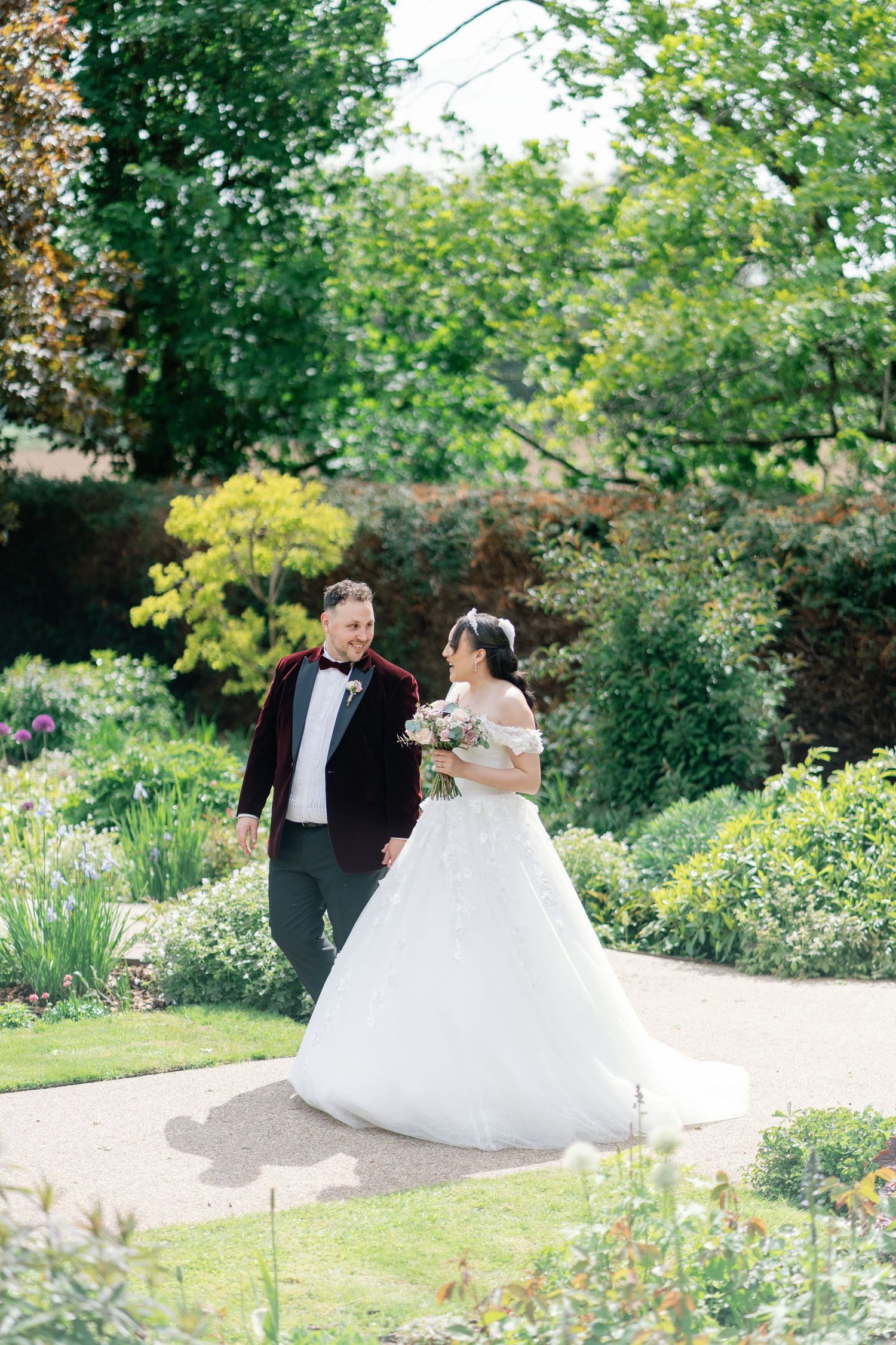 A bride in a white ball gown and groom in a burgundy tuxedo walk hand in hand through a lush garden.