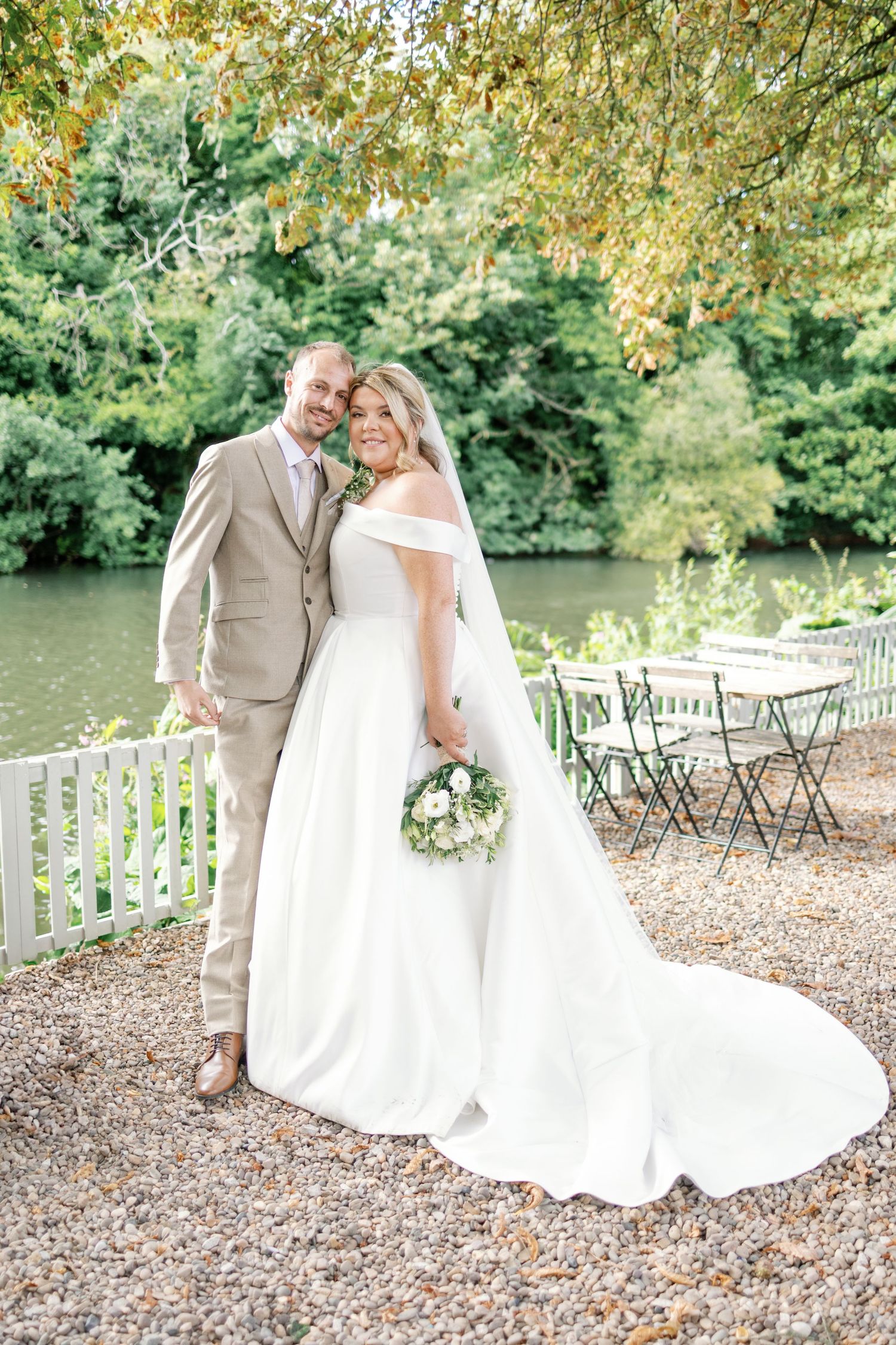 A wedding couple poses beside a tranquil pond in autumn, with scattered chairs and fall foliage in the background.