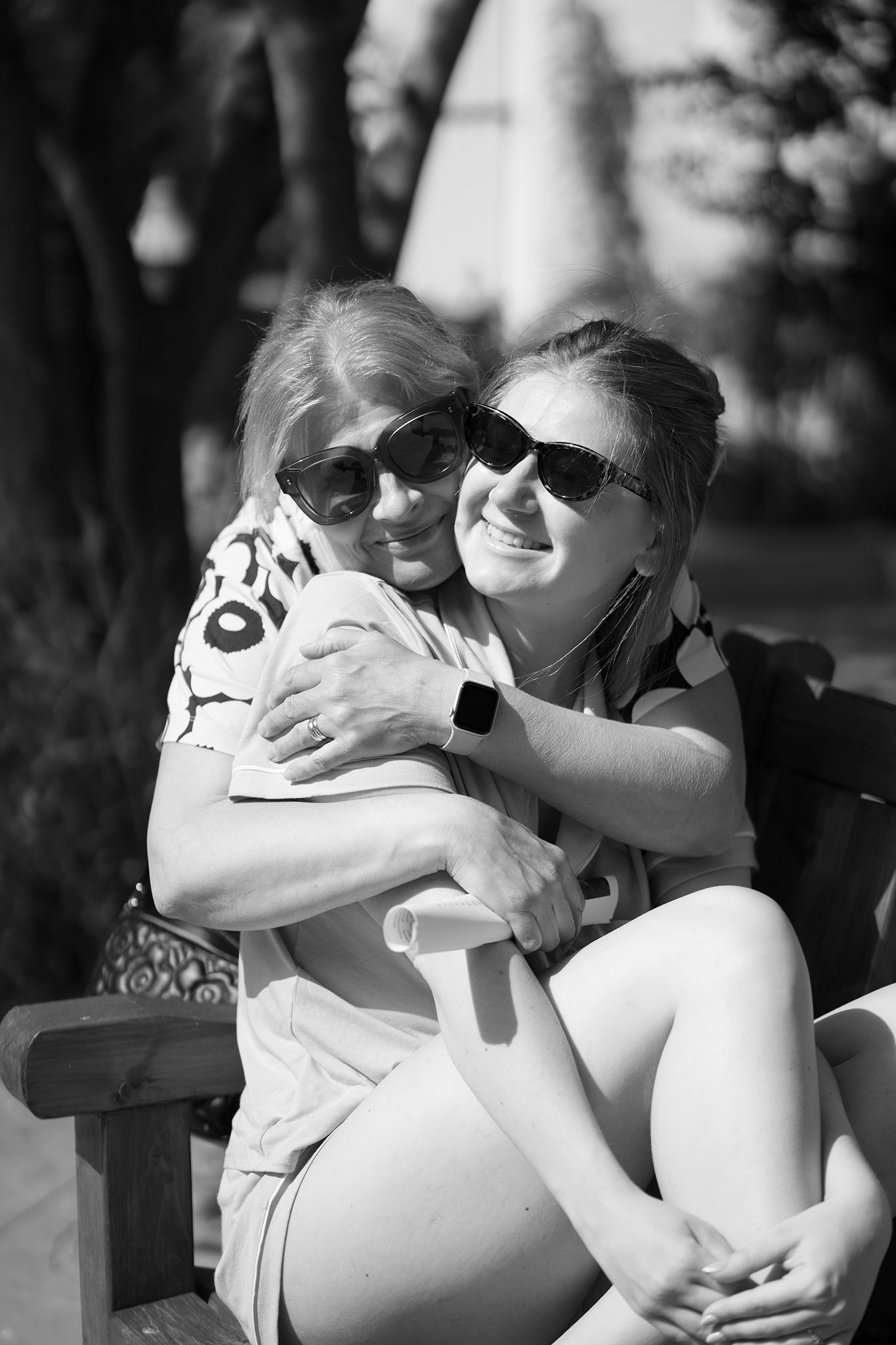 Two women hugging and smiling outdoors on a bench, wearing sunglasses in a black and white photo.