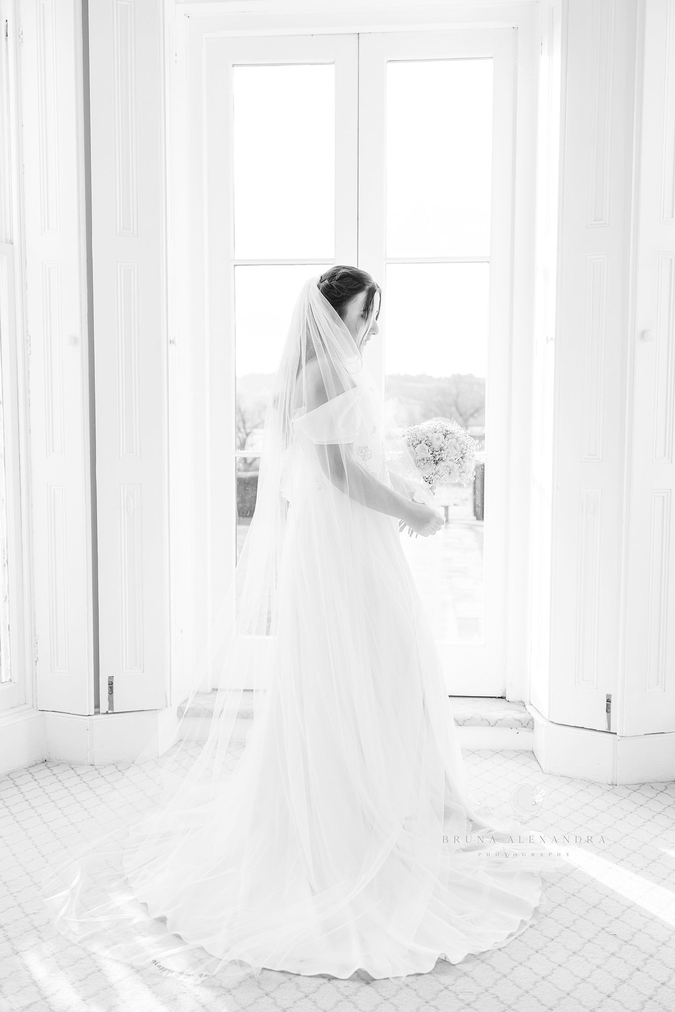 Classic black and white bridal portrait with veil and bouquet in front of bright windows.