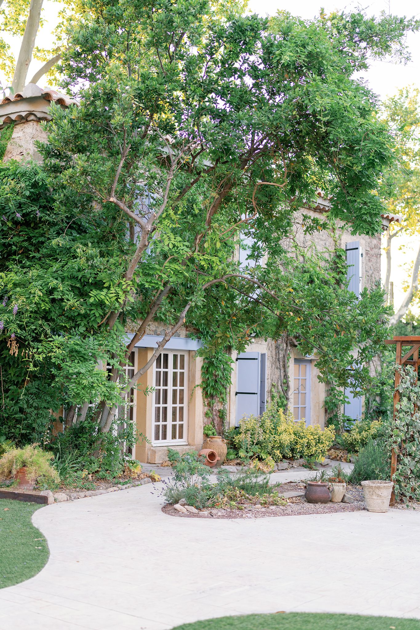 Charming stone house with blue shutters surrounded by lush green trees and manicured garden beds in a serene outdoor setting.