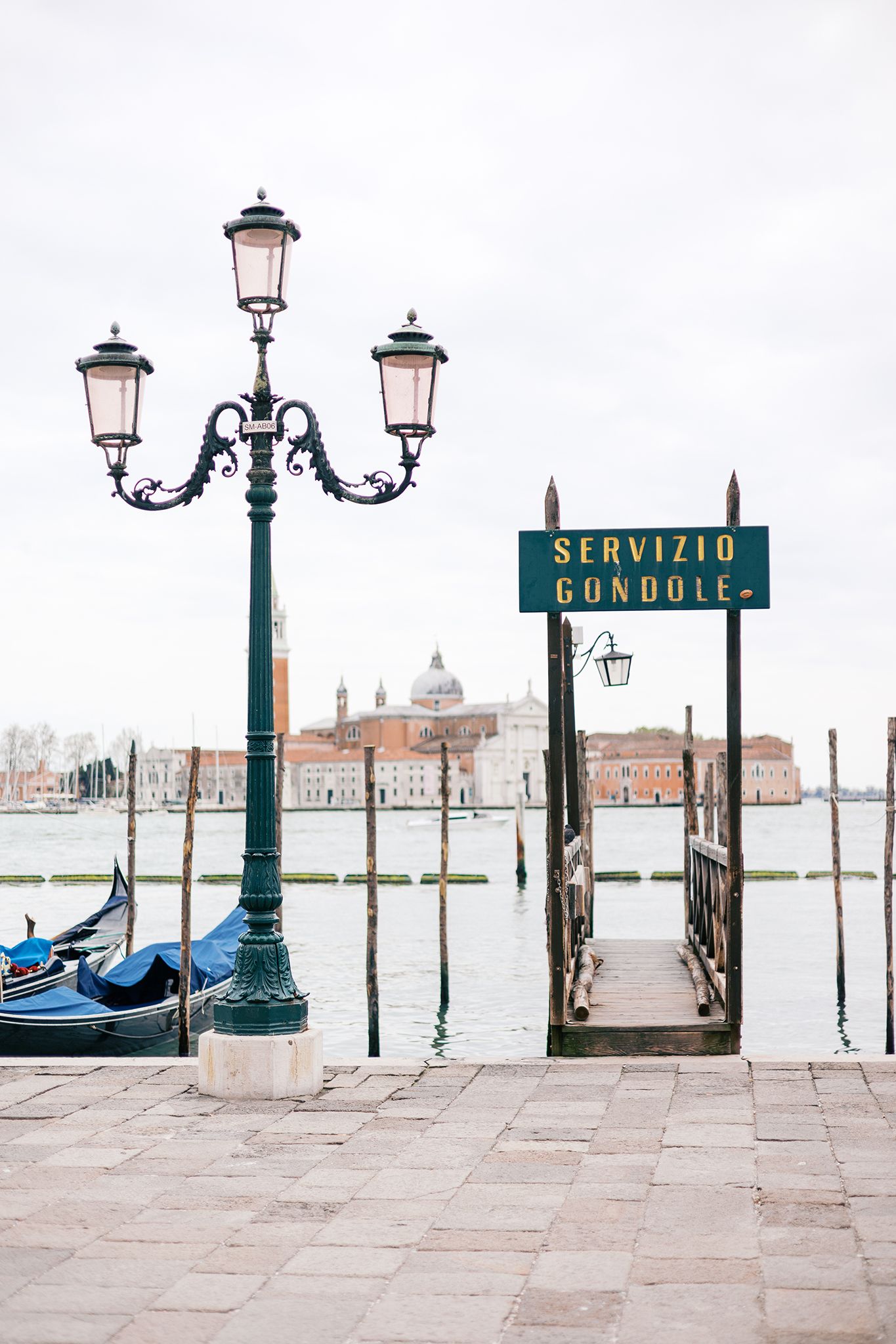 Traditional green lamp post and gondola service sign along Venice canal with San Giorgio Maggiore church in background.