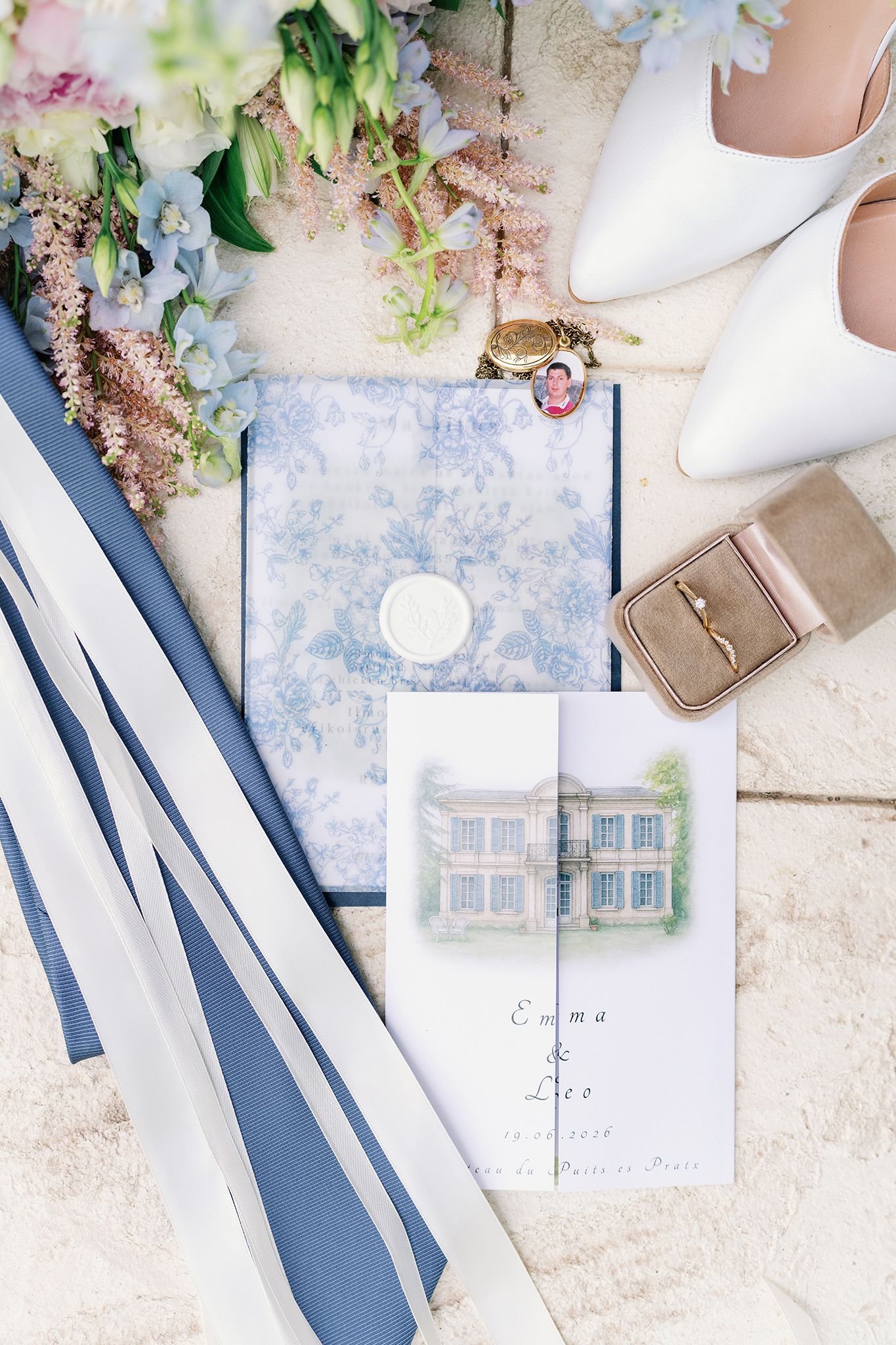 Elegant wedding flat lay with blue floral stationery, ivory heels, ring box, bouquet, and personalized invitation for Emma and Leo.