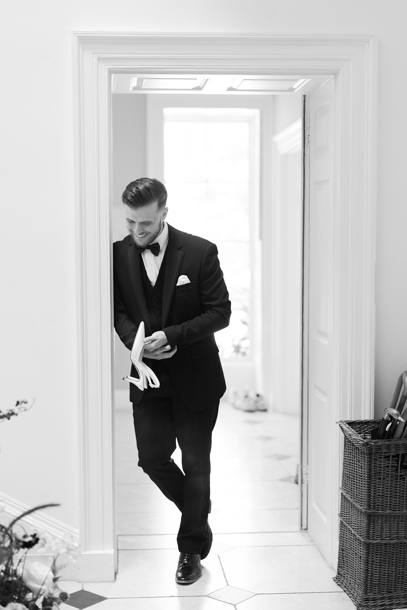 Elegant groom in black tuxedo with bow tie adjusting cufflinks while standing in a bright white doorway.