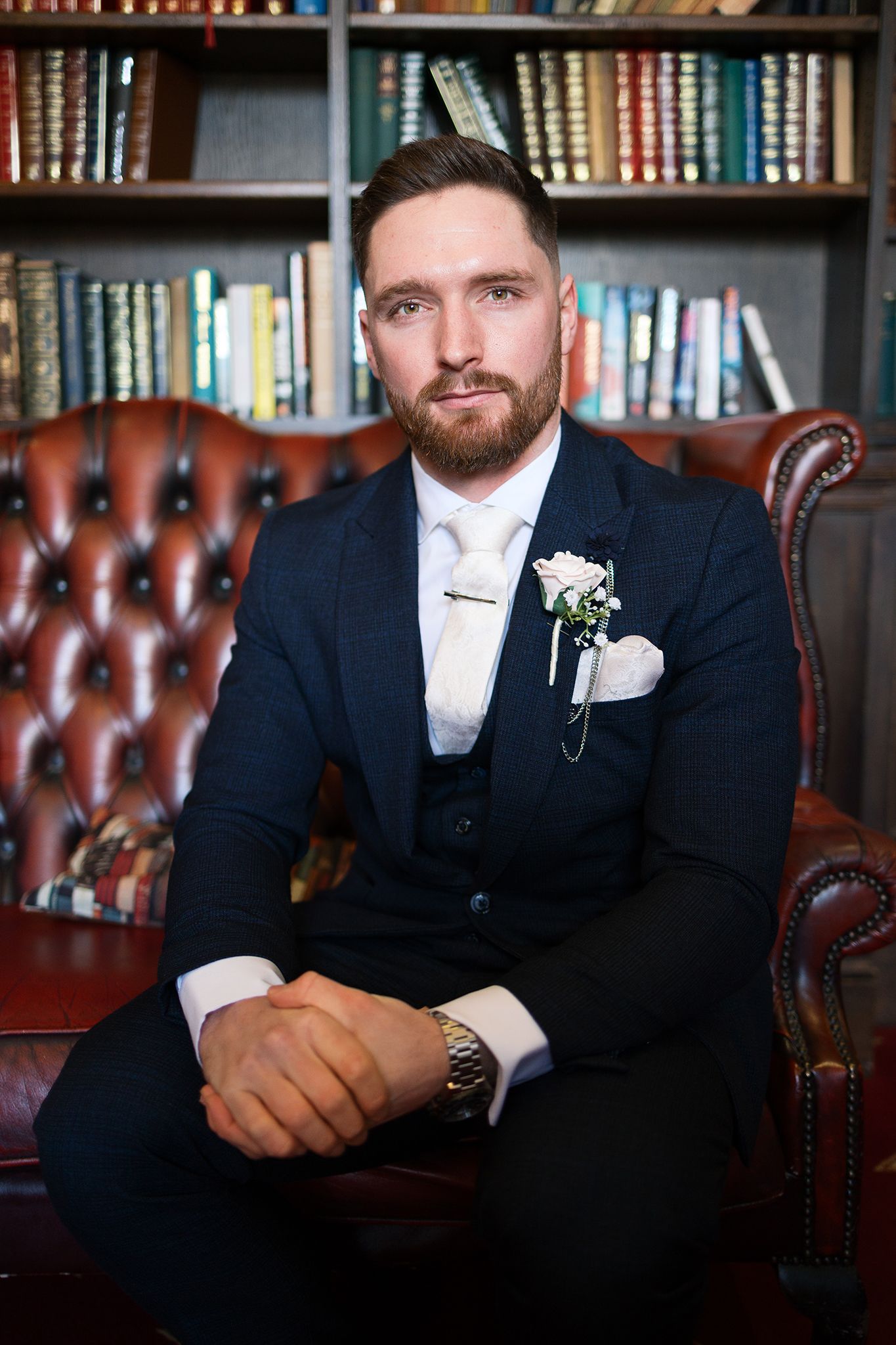 A groom in a navy suit and white boutonnière sits on a brown leather chair in front of a bookshelf.