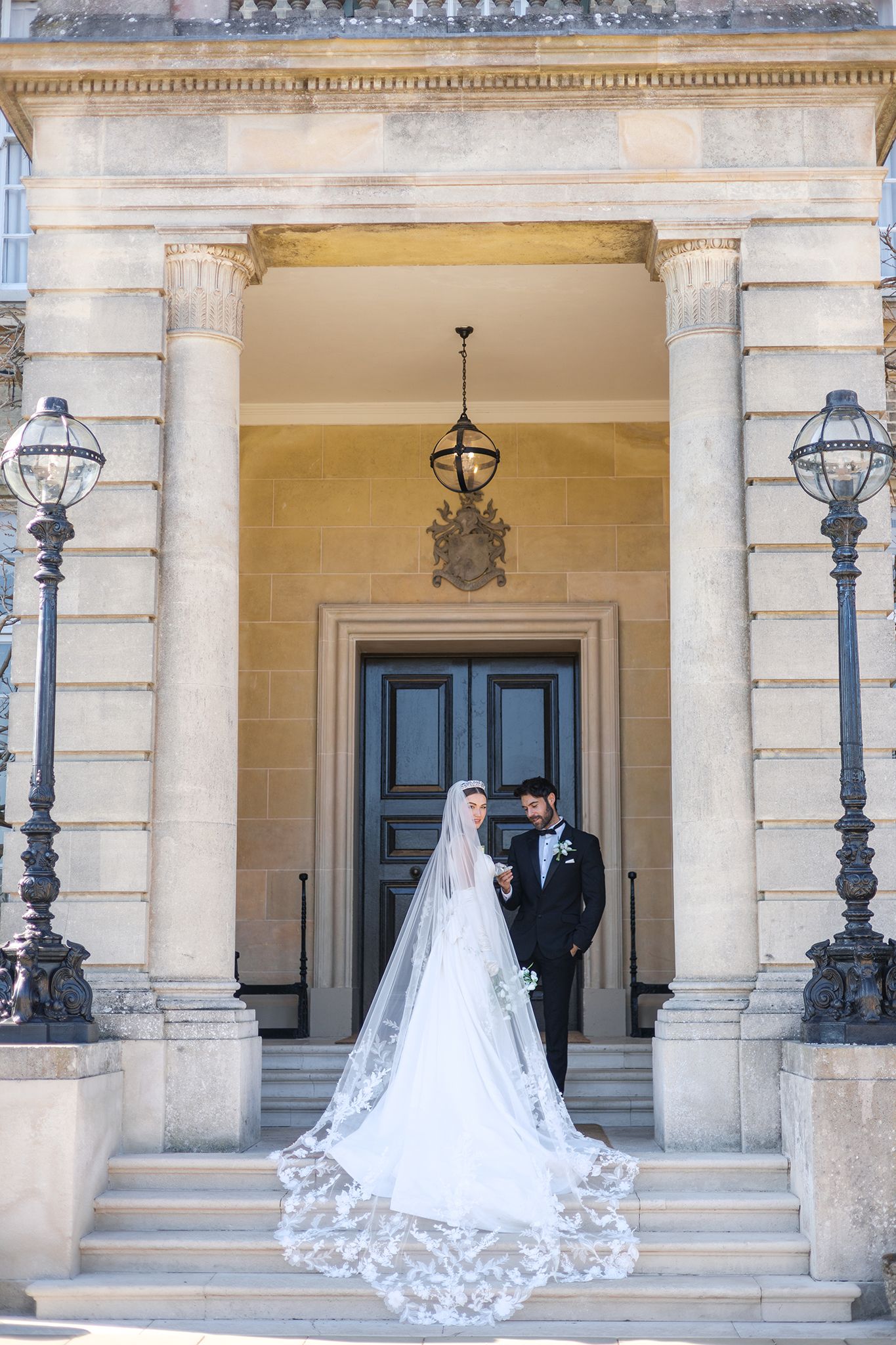 Bride and groom pose on grand stone steps of elegant venue, cathedral veil trailing, between ornate columns.