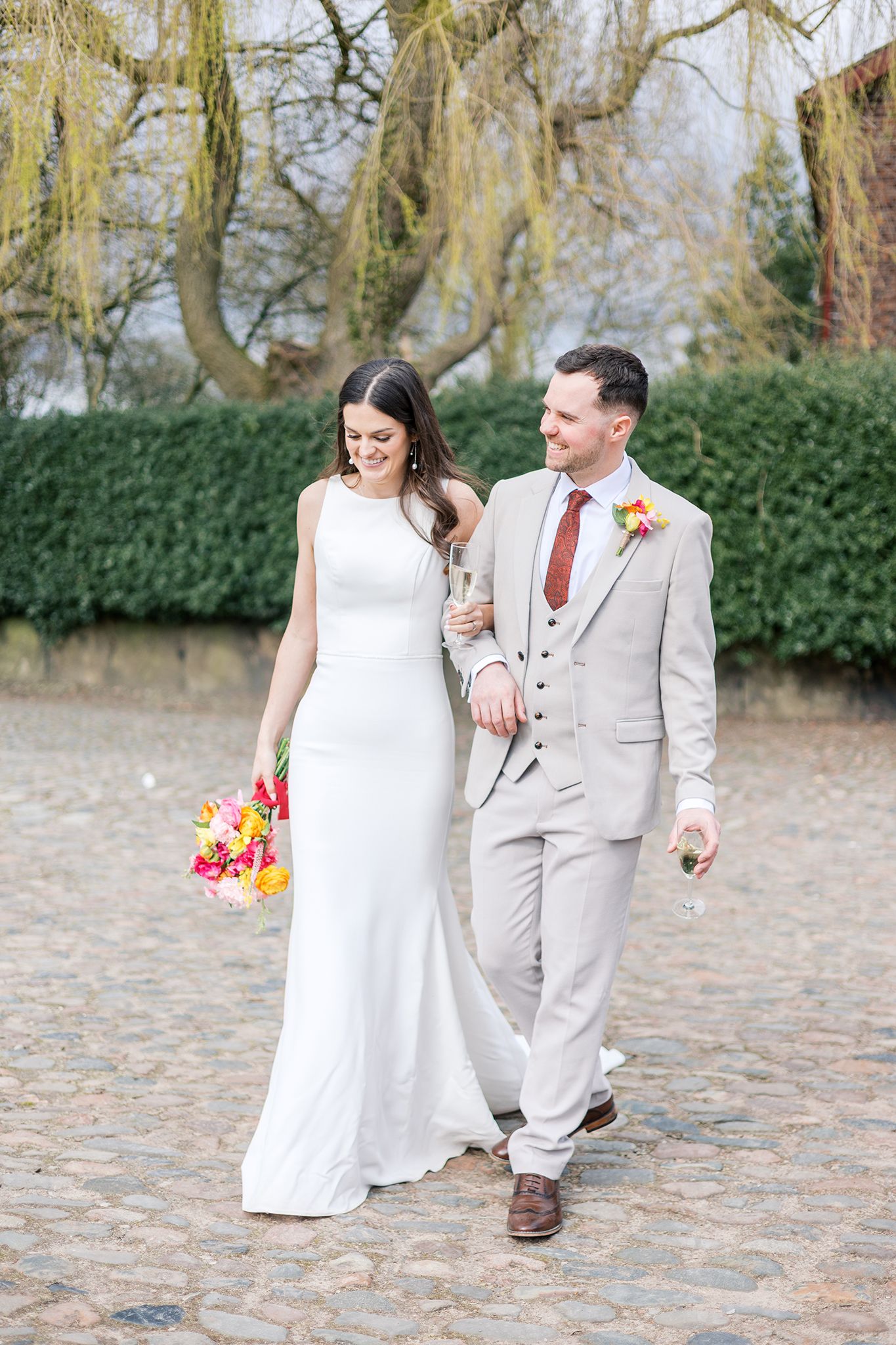 A couple in wedding attire walks together on a cobblestone path with willow trees and greenery in the background.