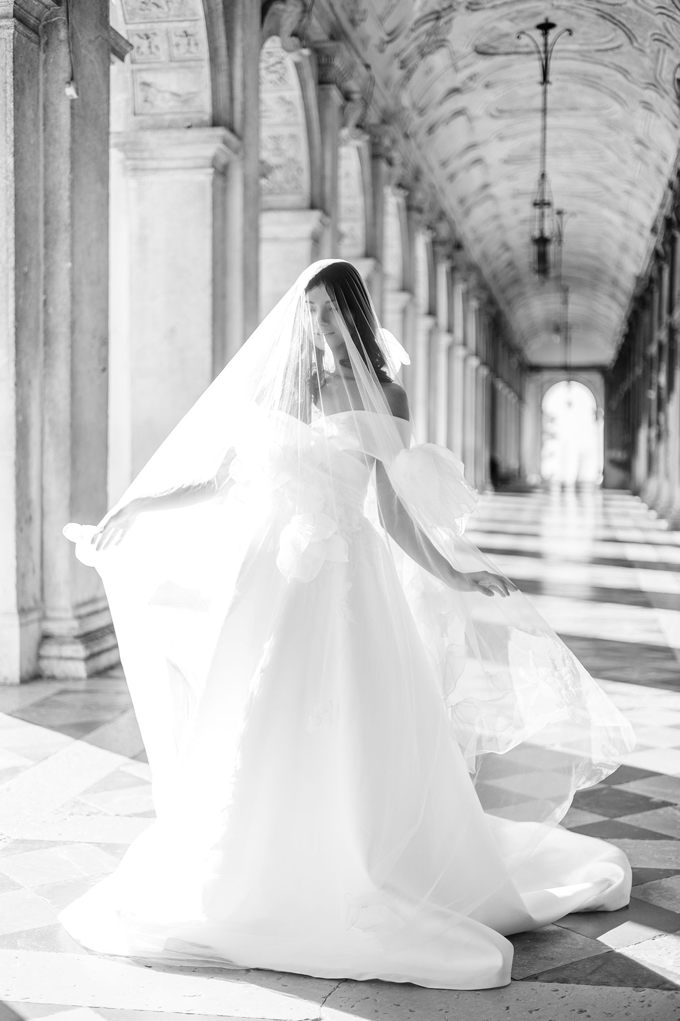 A bride in a flowing white wedding dress and veil twirls elegantly in a grand hallway with ornate columns and vaulted ceilings.