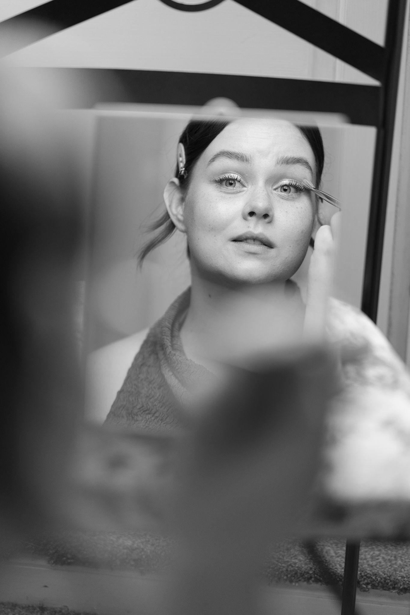 Black and white photo of a young woman applying eye makeup while looking in a mirror, with a tattoo visible on her arm.