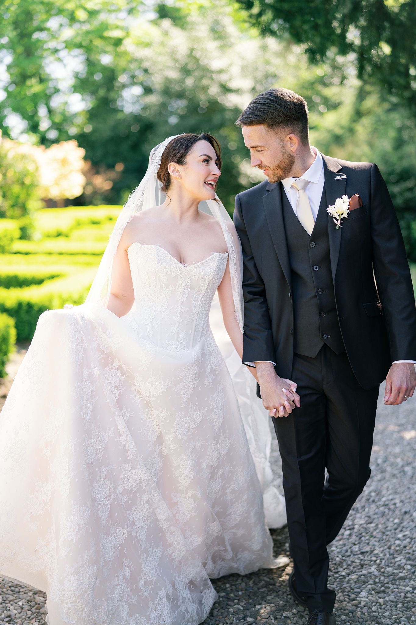 A bride and groom walk hand in hand through a sunlit garden, smiling at each other on their wedding day.