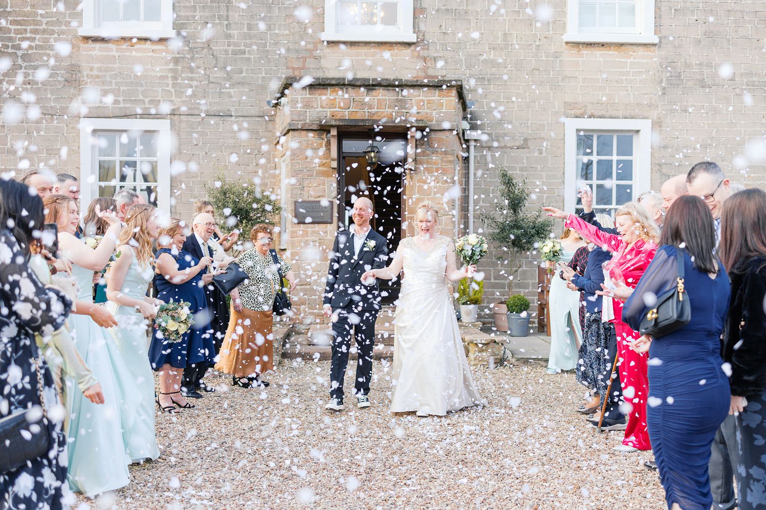 Wedding celebration outside stone building with guests throwing white confetti in the air during joyful outdoor ceremony.
