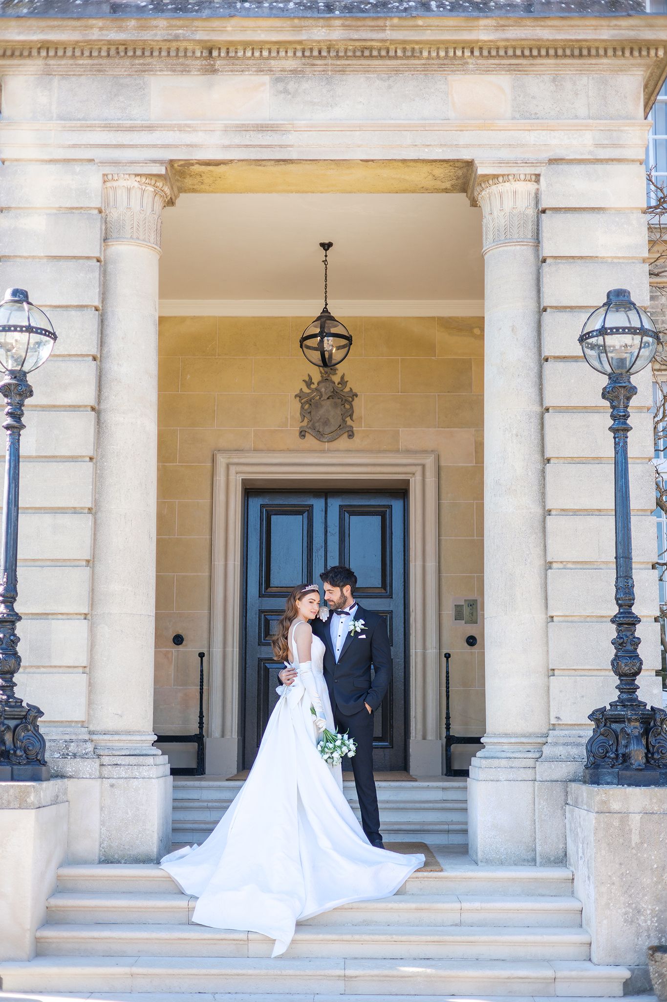 A couple in wedding attire embrace on an elegant stone portico with ornate lamp posts and a grand blue door.