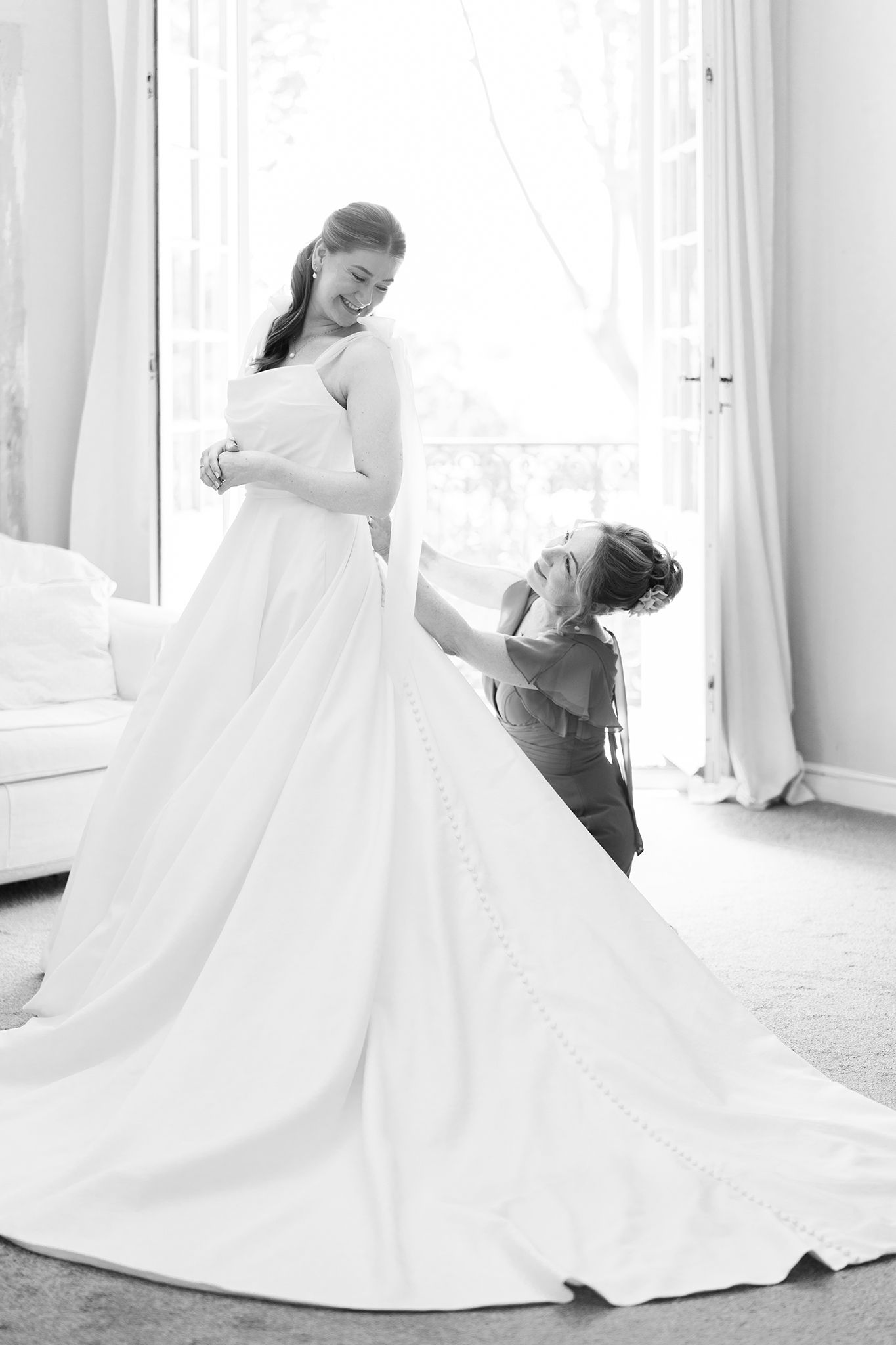 Black and white photo of a bride in a flowing white gown being helped by a bridesmaid in an elegant bright room.