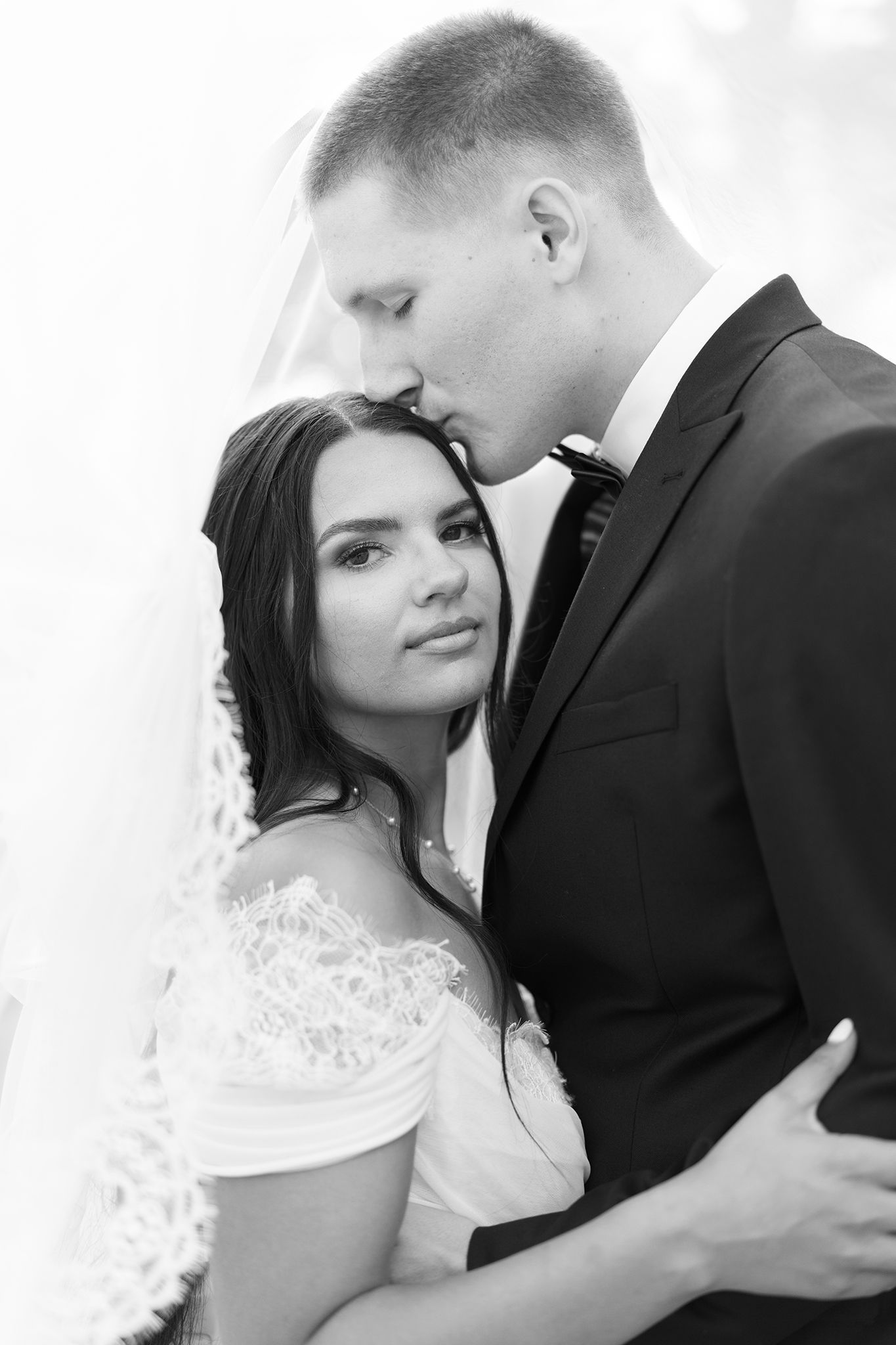 Black and white wedding photo of bride and groom embracing, groom kissing bride on forehead.