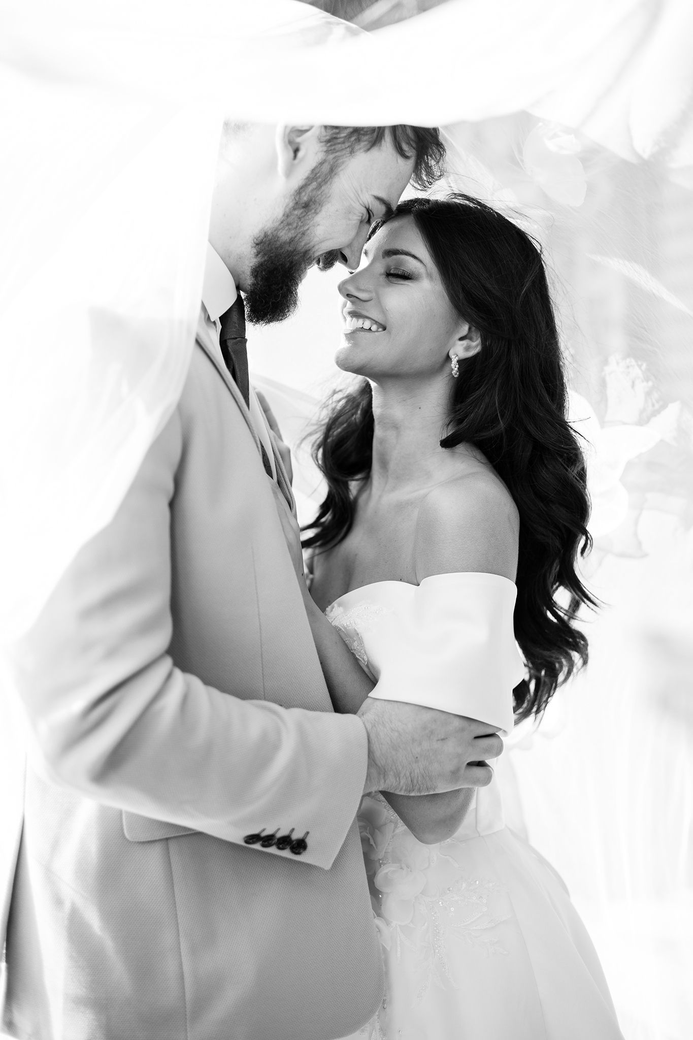 Romantic black and white wedding portrait of a couple sharing an intimate moment under flowing fabric.