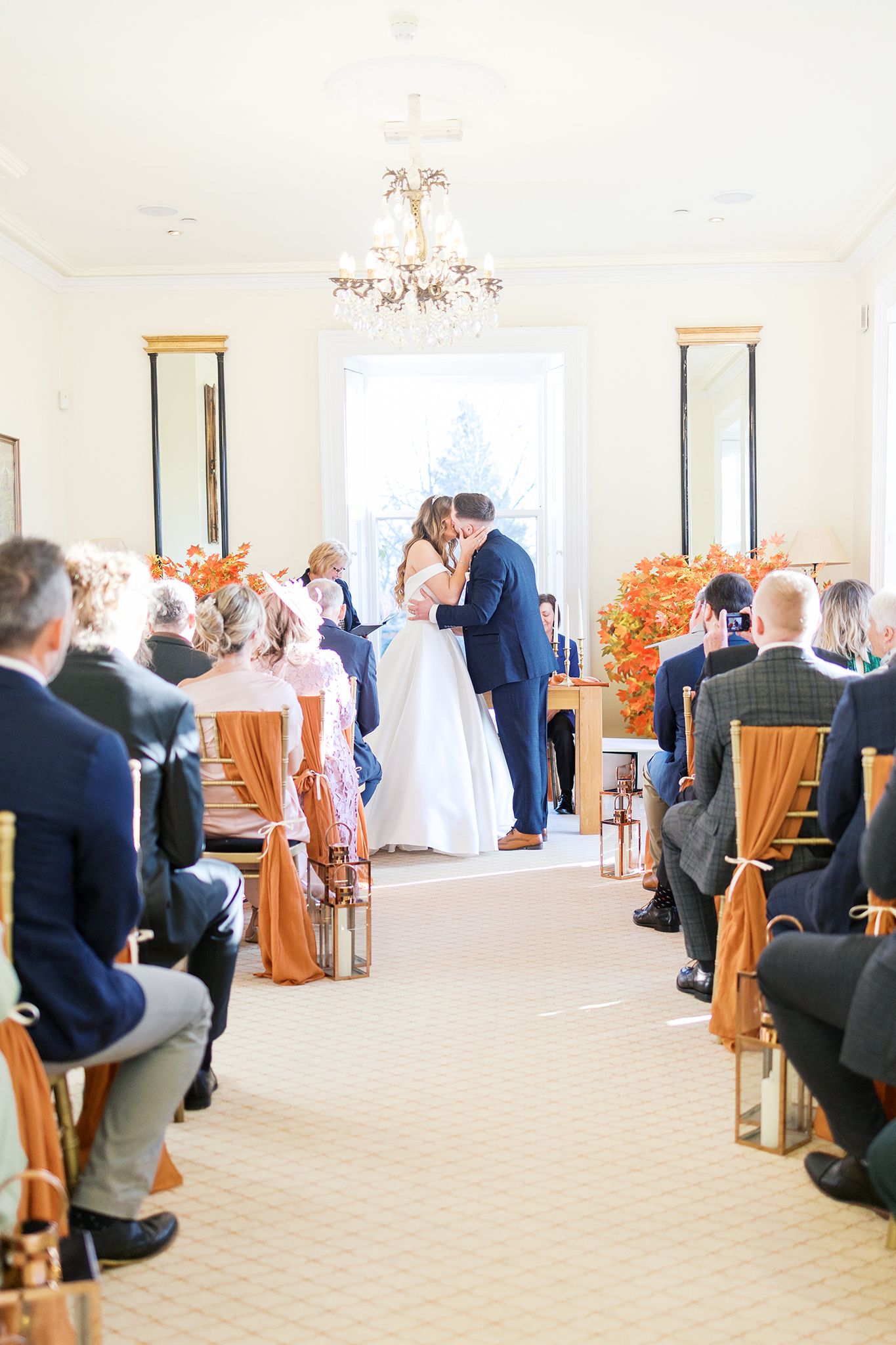 A wedding ceremony in a bright white room with guests seated in wooden chairs and orange floral arrangements.