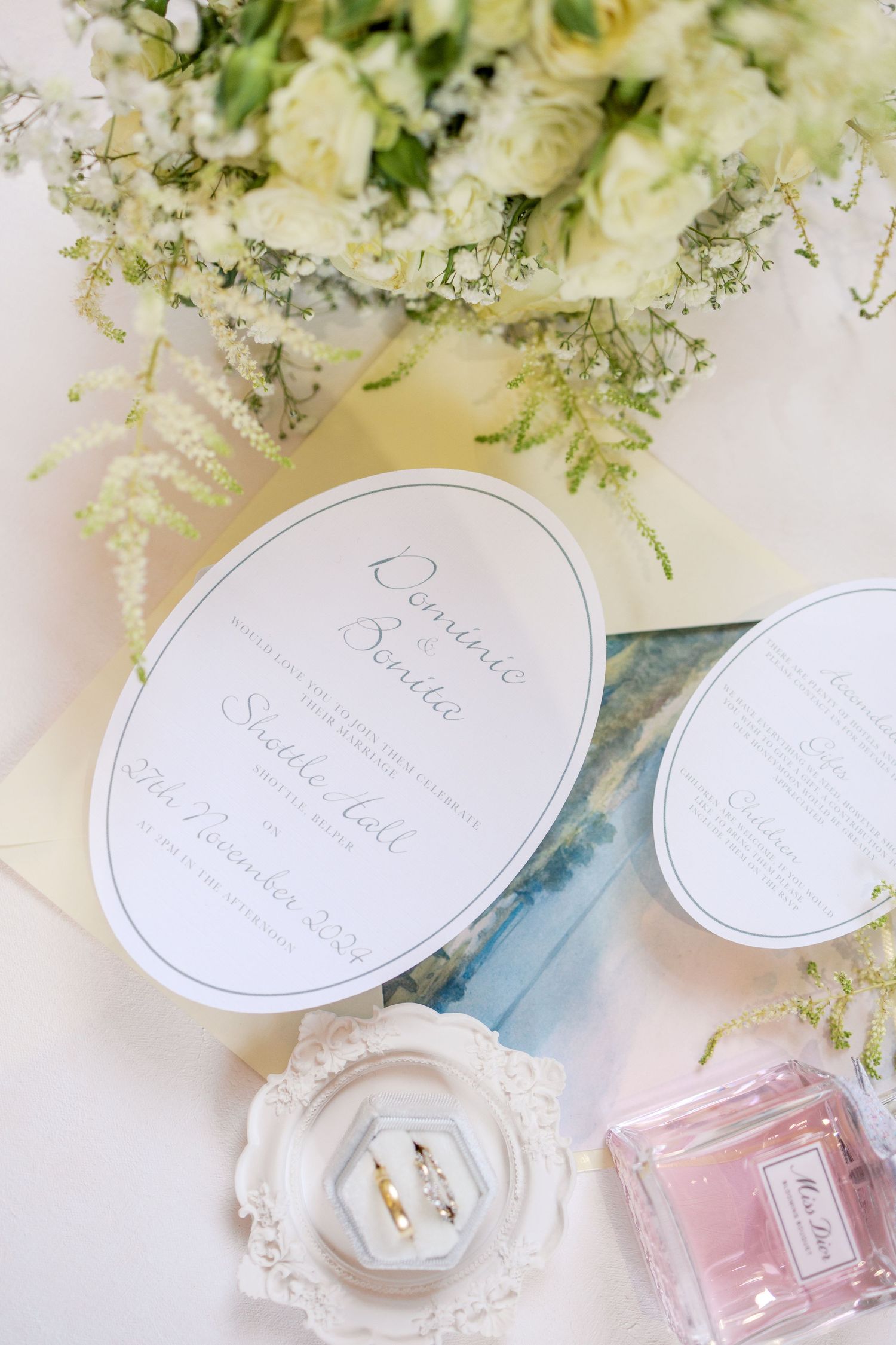Wedding details featuring white invitation cards, flowers, and perfume bottle on a light surface.
