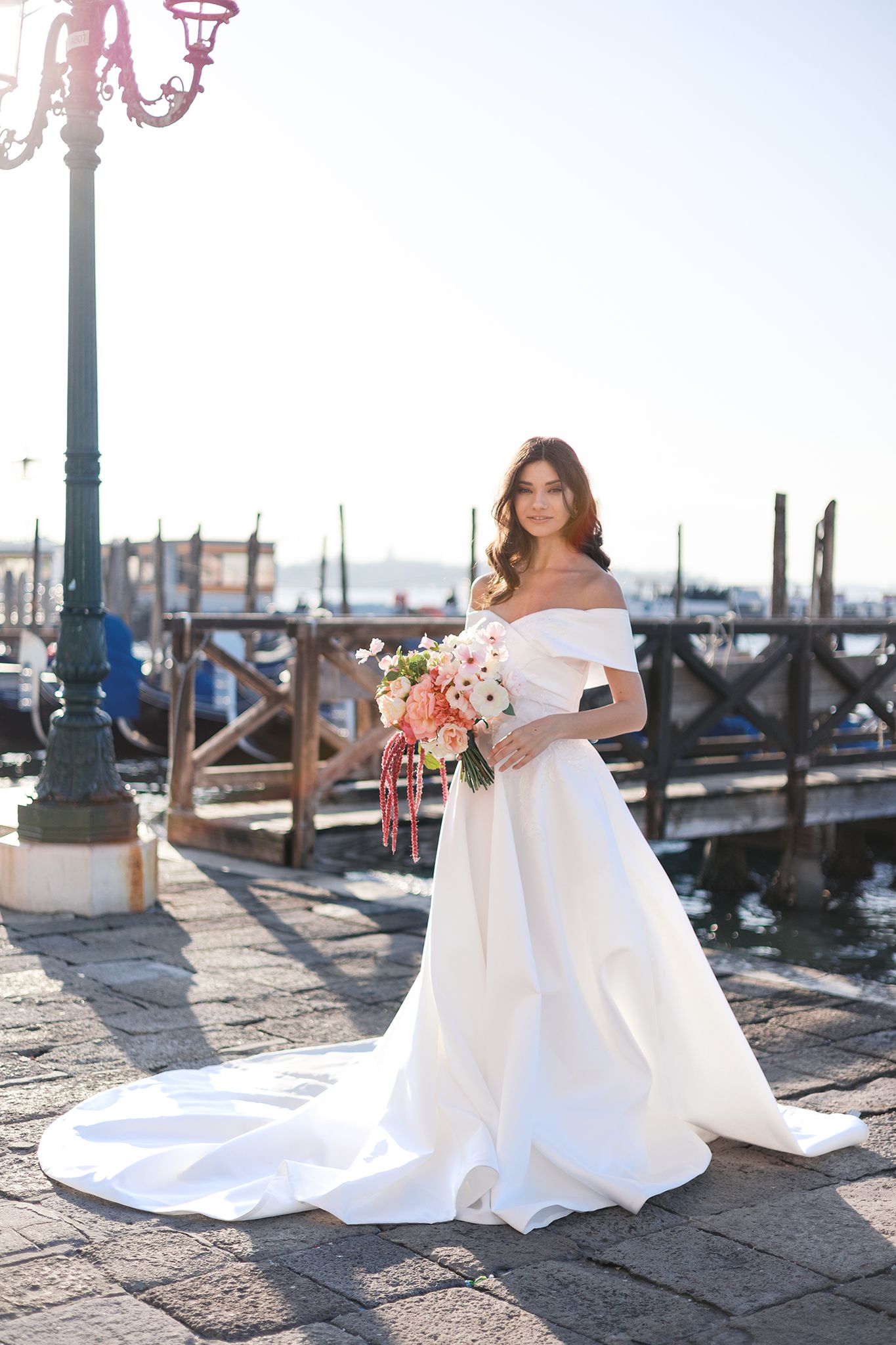 Elegant off-shoulder white wedding gown with long train photographed at a scenic waterfront dock in Venice.