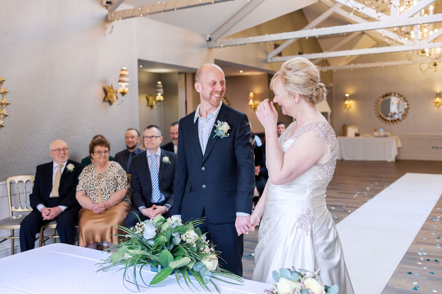 Couple at romantic indoor wedding ceremony with white decor and floral arrangements near a decorative table in barn venue.
