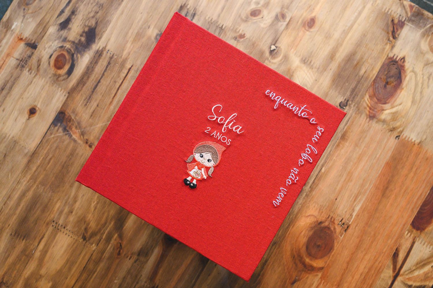 A red photo album or scrapbook with decorative text lies on a wooden surface with visible knots and grain patterns.