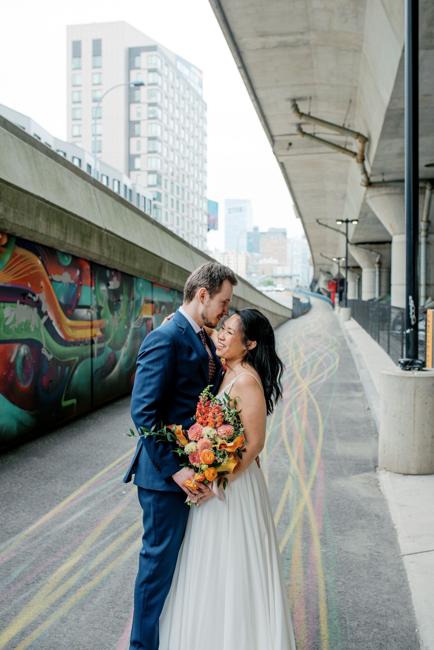 Modern wedding couple embraces under urban concrete overpass with colorful graffiti murals and architectural lines.