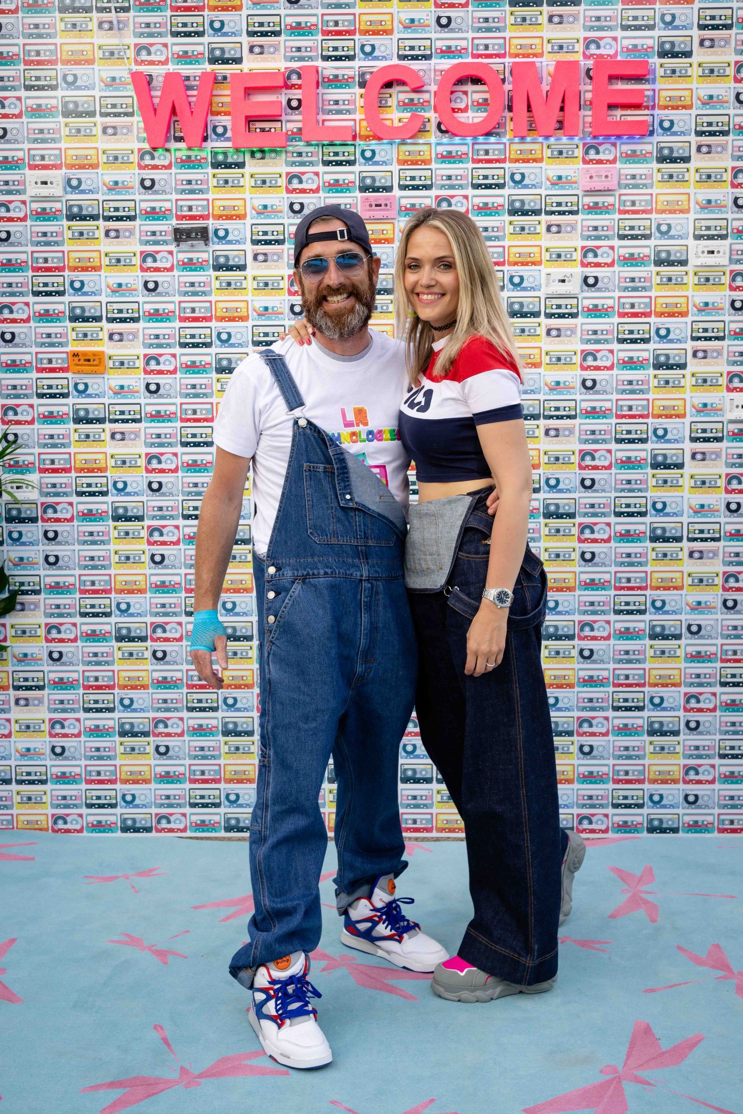 People wearing denim overalls pose casually against wall covered in vintage cassette tape artwork.
