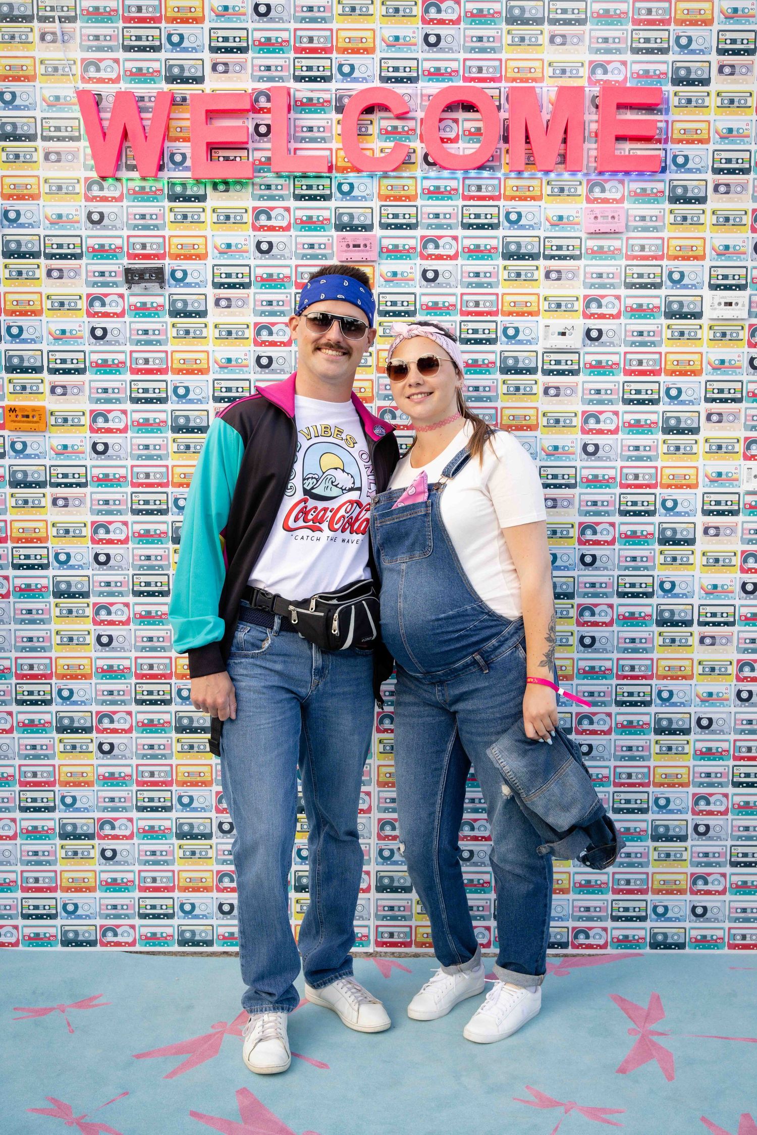 Two people pose in casual denim outfits against a colorful welcome wall with retro cassette tape design.