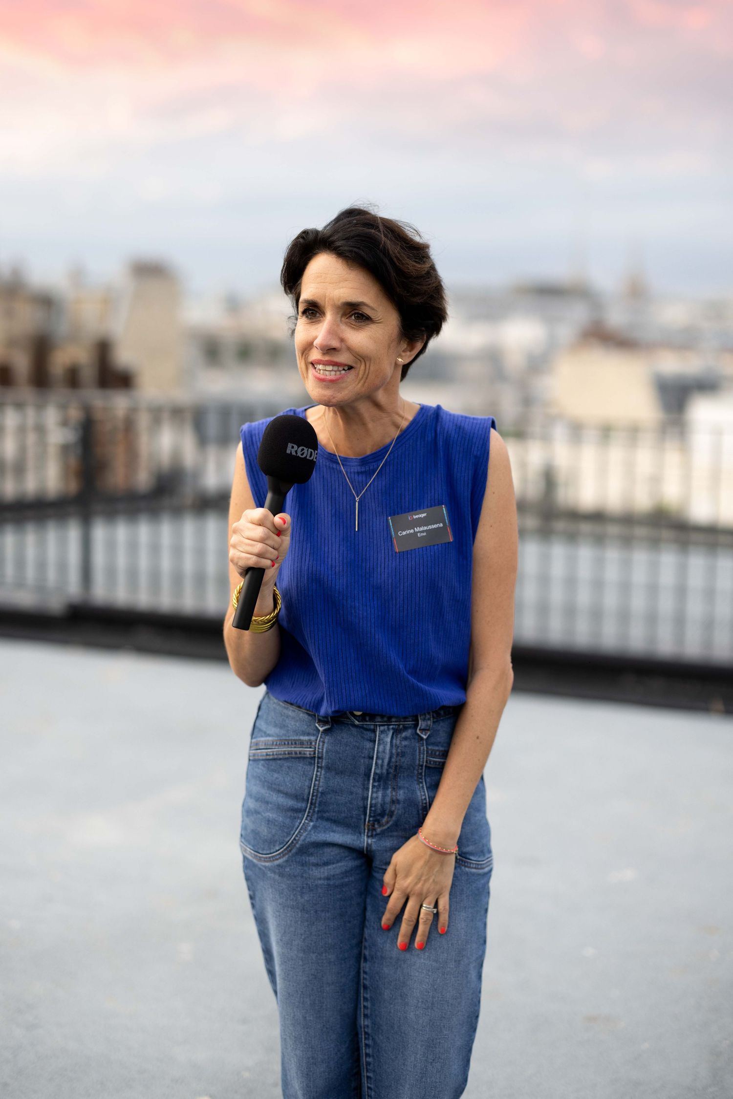 Speaker in blue sleeveless top presenting with microphone on rooftop venue.