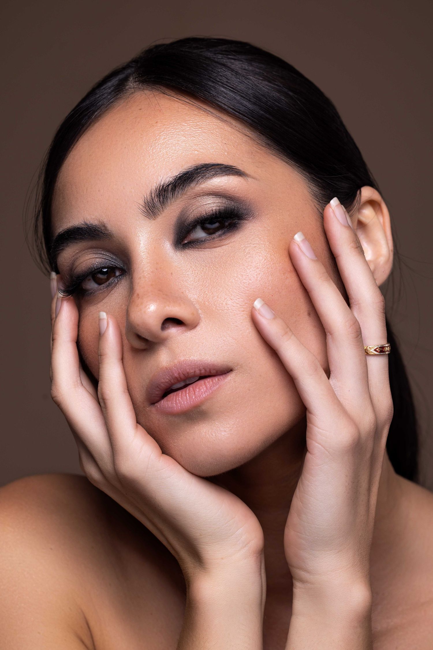 Gros plan beauté montrant un maquillage lumineux et des mains encadrant le visage sur fond moka.