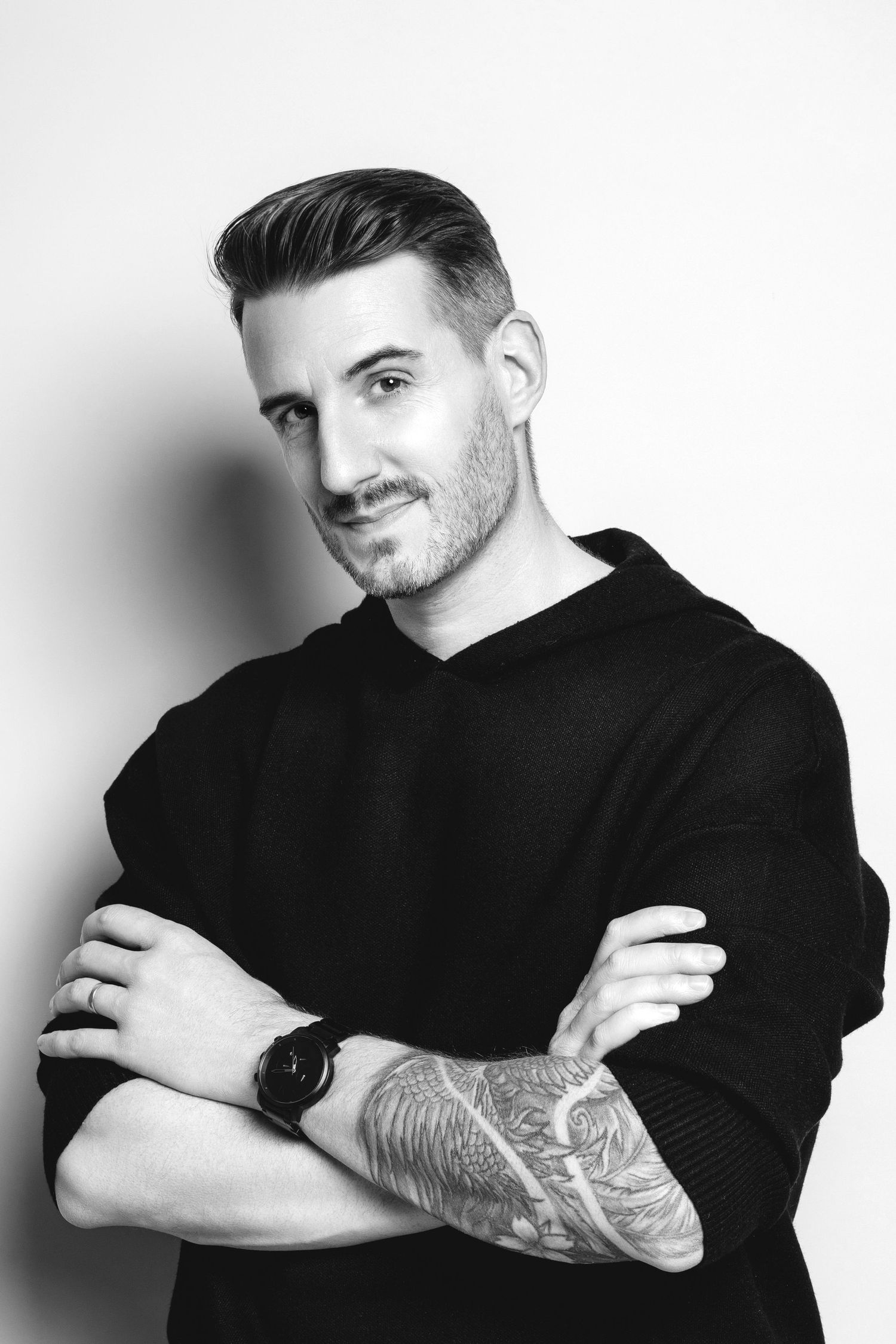 Black and white portrait of a tattooed man in a black hoodie with arms crossed, smiling confidently.