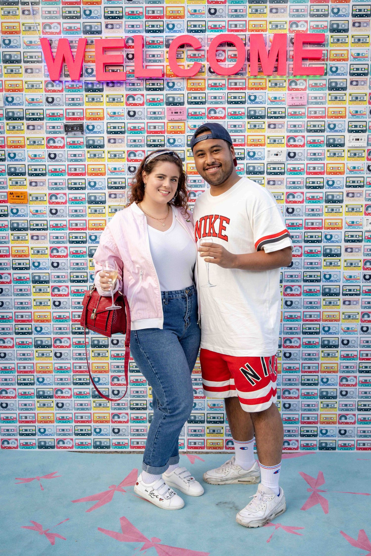 Couple in casual streetwear poses together against a wall decorated with colorful cassette tapes.