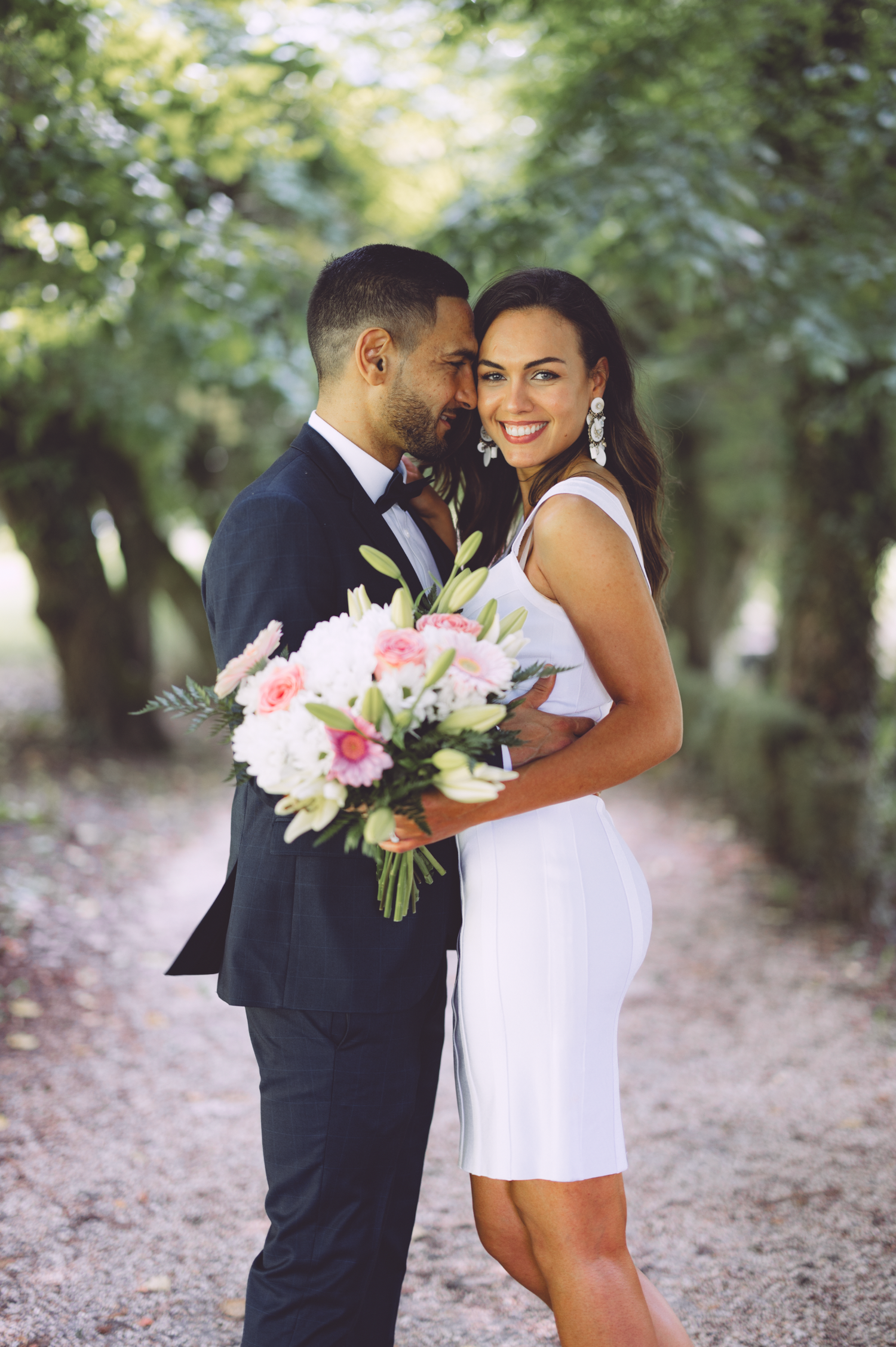 A couple shares a romantic moment on a tree-lined path while holding a pink and white floral bouquet.