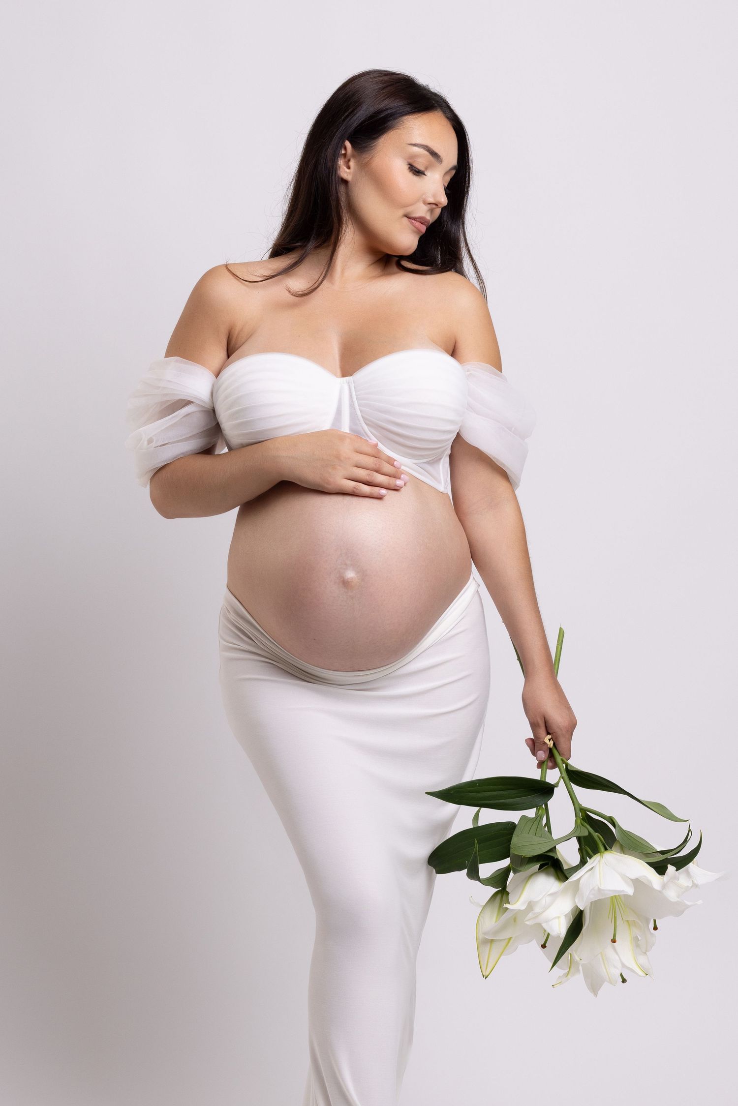 A maternity photoshoot series featuring an elegant white dress and white lily flowers against a light background.