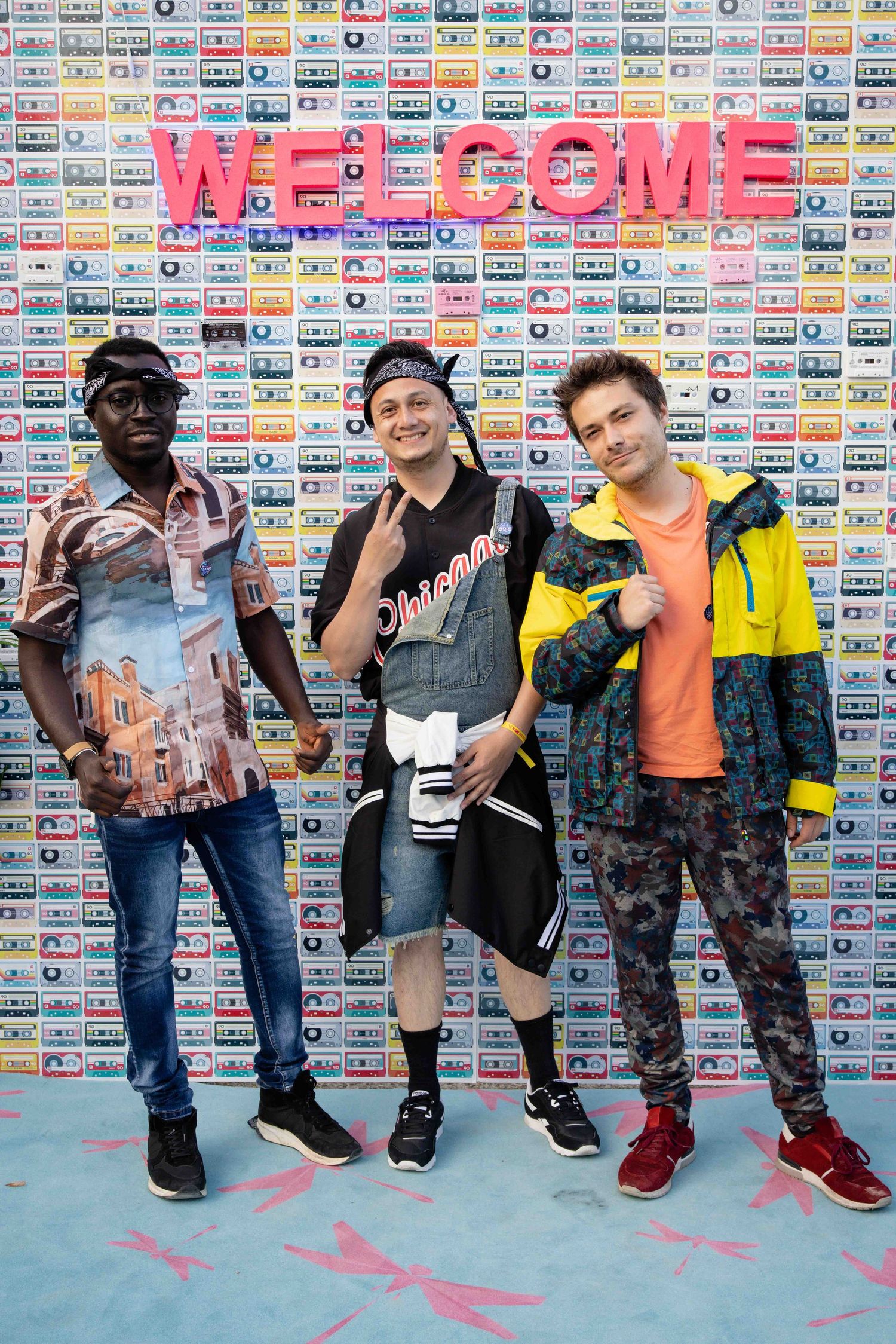 Three friends strike poses in front of a decorative welcome wall with vintage cassette tape background.
