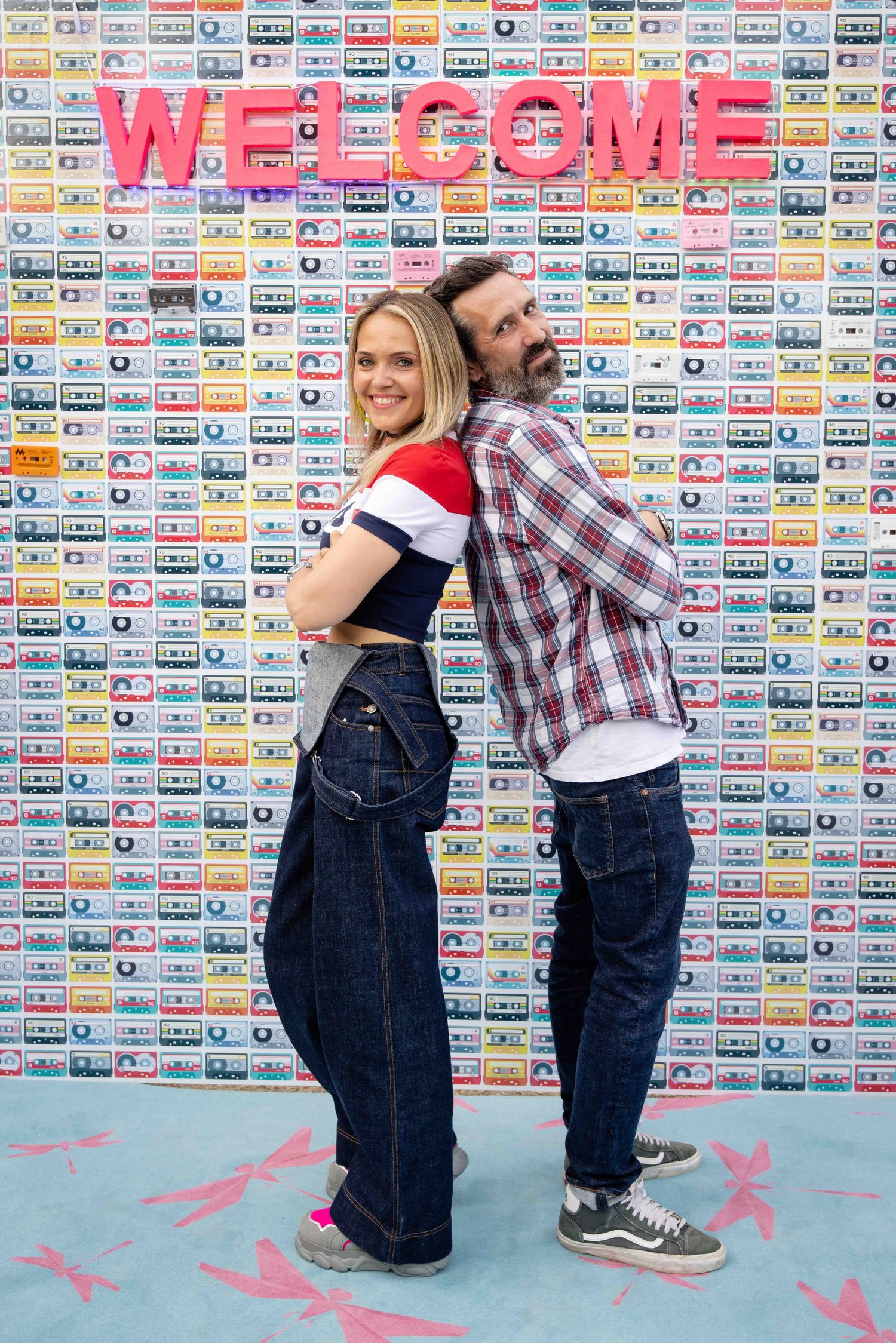 People in retro denim outfits strike playful poses against wall covered in vintage cassette tapes.
