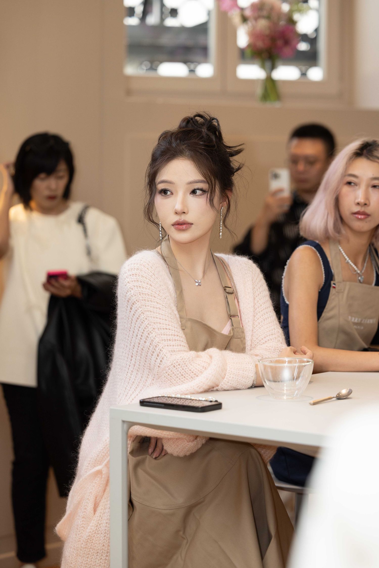 Attendee in pink sweater and beige apron seated at white table during gathering.