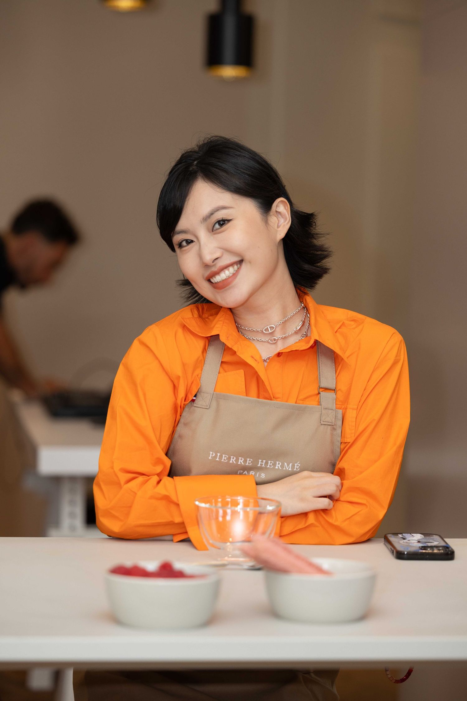 Person in bright orange apron smiling while seated at white counter during event.