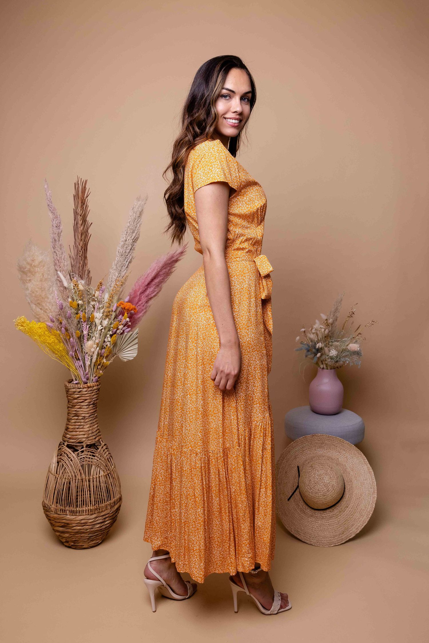 Une femme en robe fleurie jaune pose avec des compositions de fleurs séchées et un chapeau de paille.