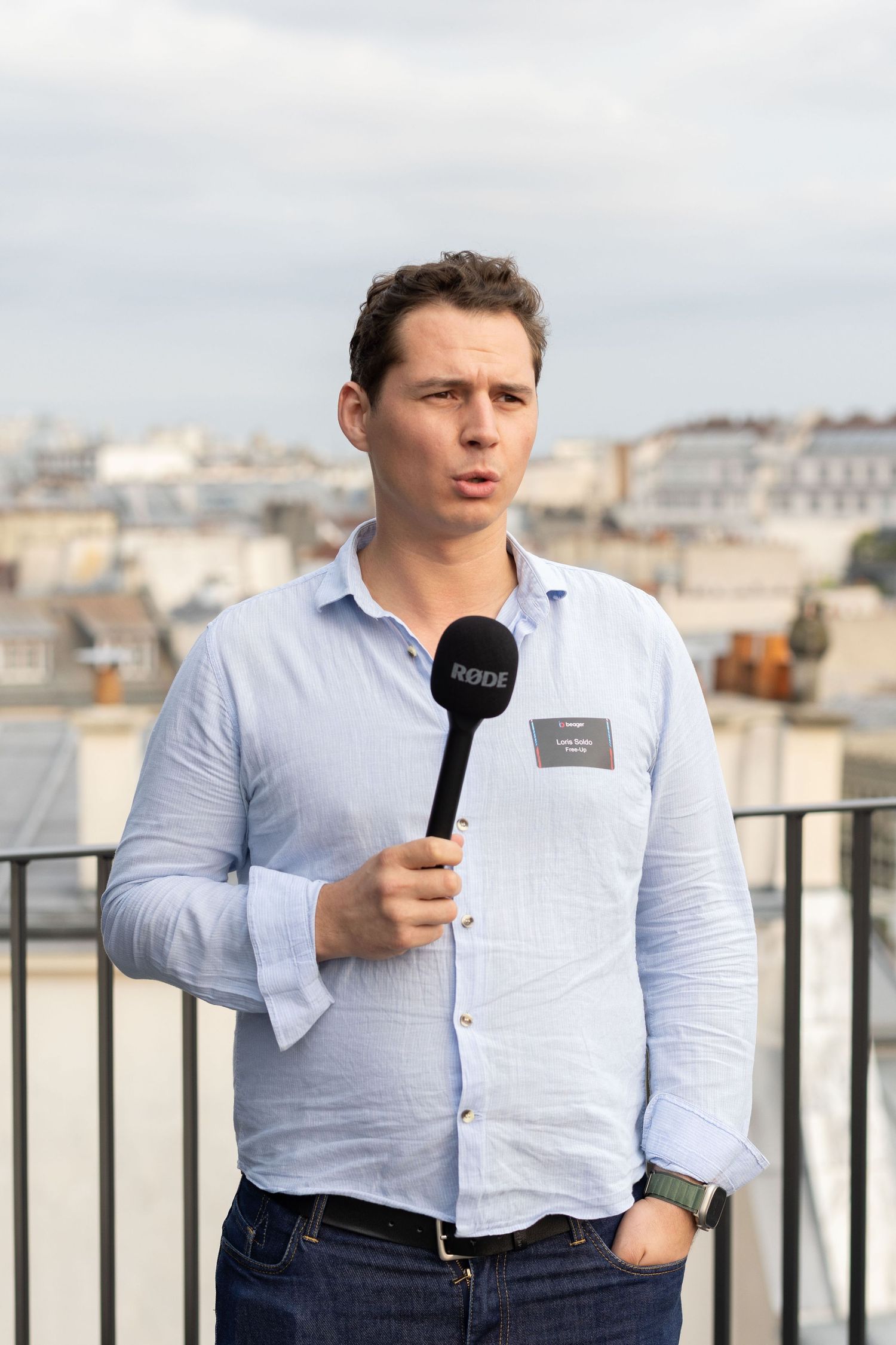 Person in light blue shirt holding microphone on rooftop terrace with city backdrop.