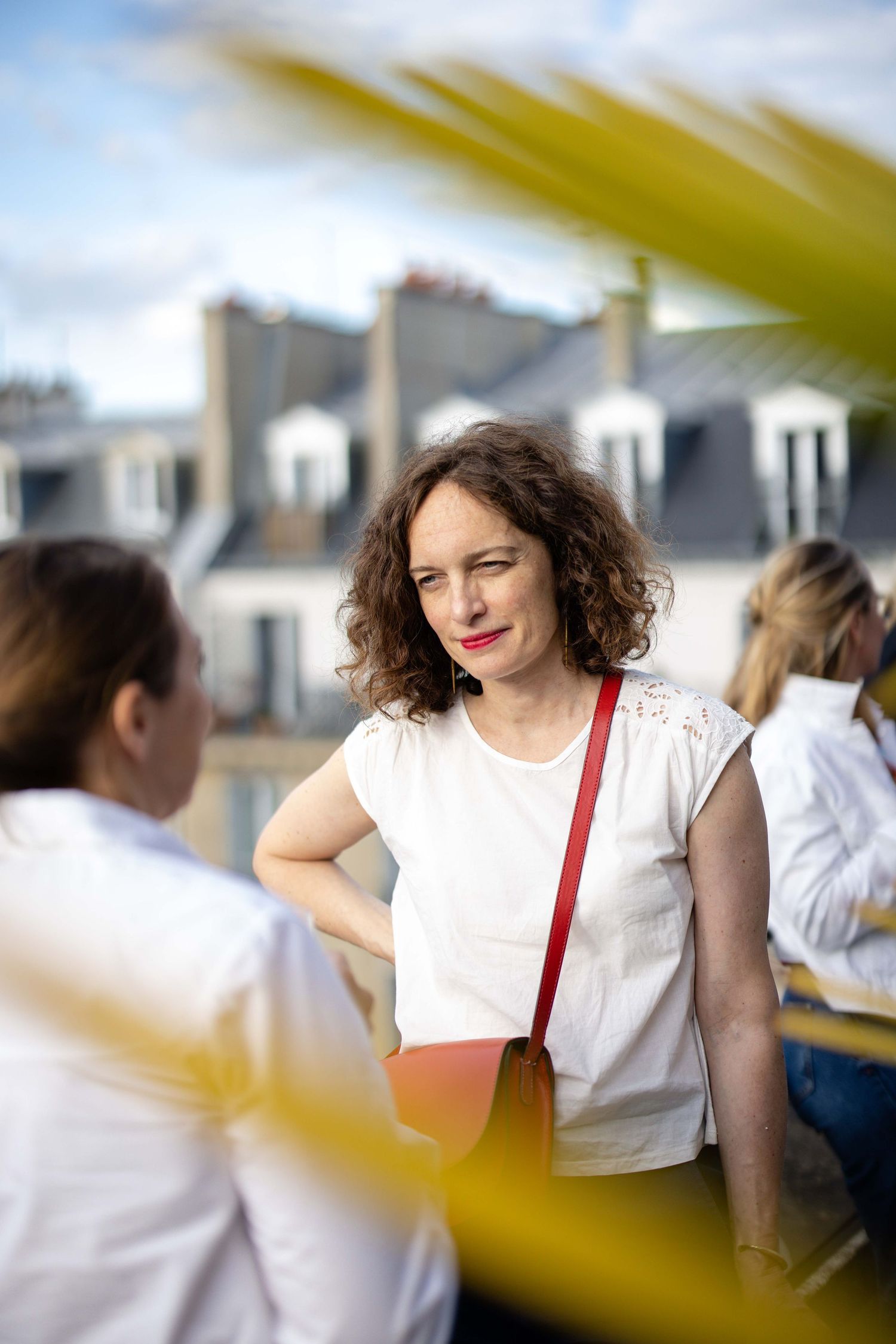 A person with curly hair wearing a white shirt and red strap smiles during a social gathering.