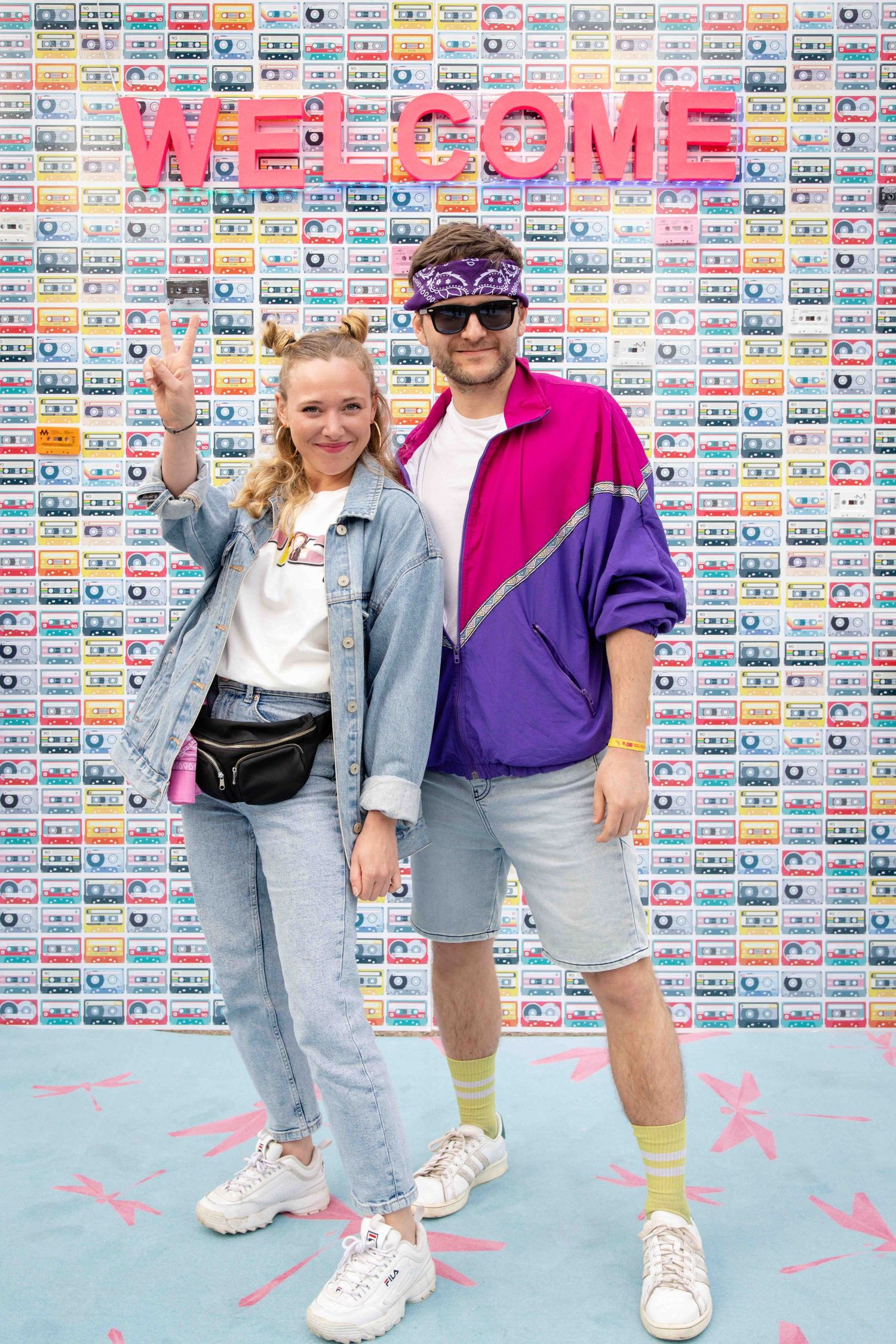 Group poses against wall decorated with colorful cassette tapes and pink welcome sign wearing retro 90s fashion.