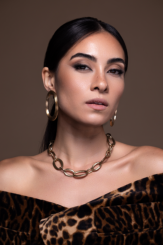 Woman in leopard print top wearing gold hoop earrings and chain necklace poses against brown background.