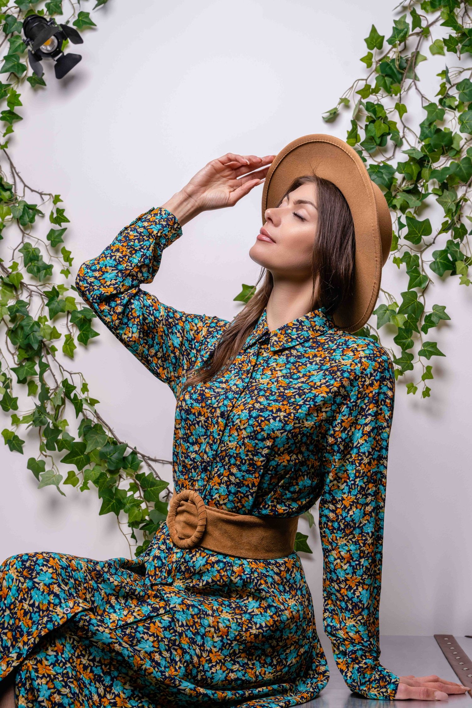 Modèle en robe fleurie et chapeau marron pose avec un décor de lierre grimpant.