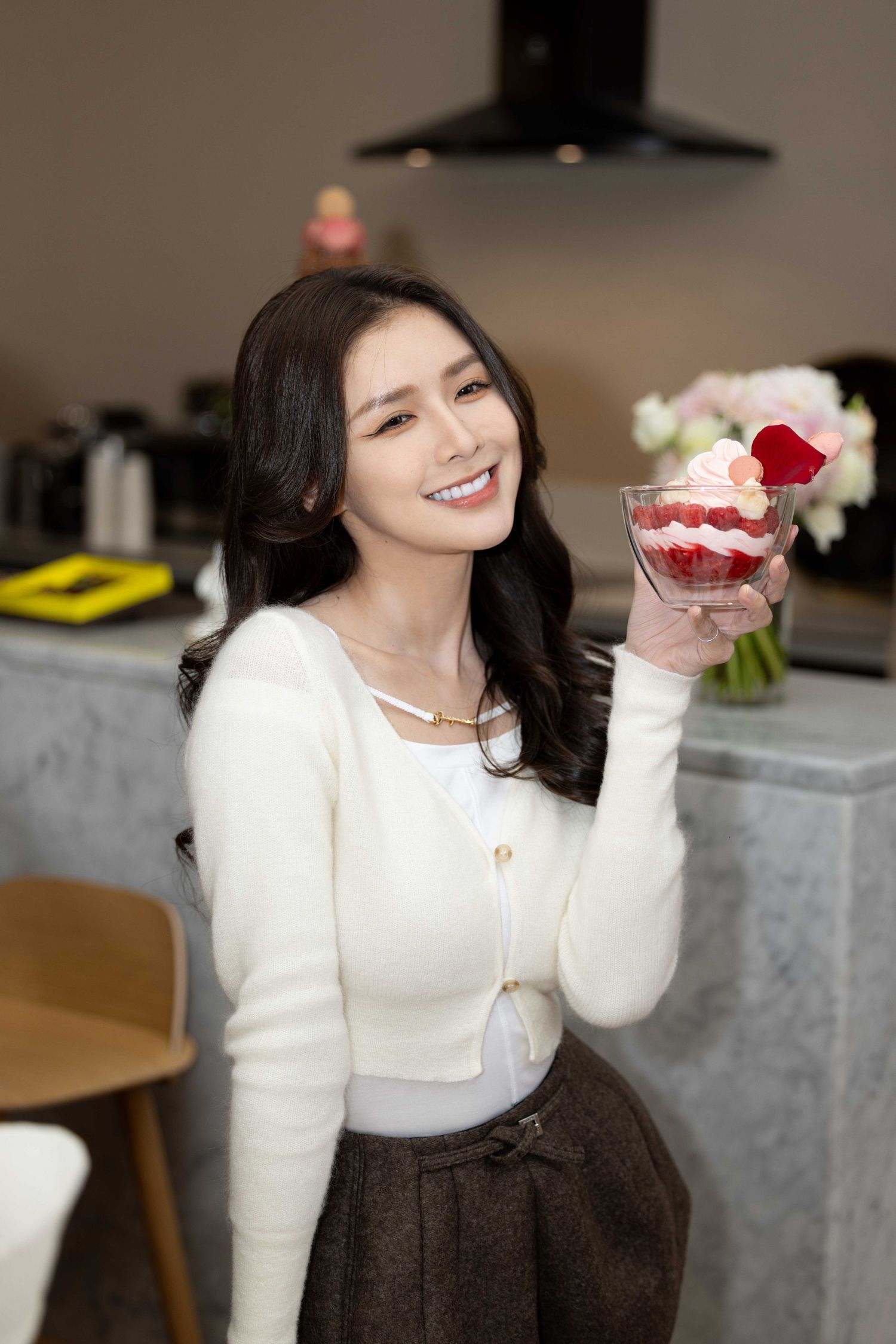 Woman in white sweater and dark skirt holds pink dessert while smiling in cozy indoor setting.
